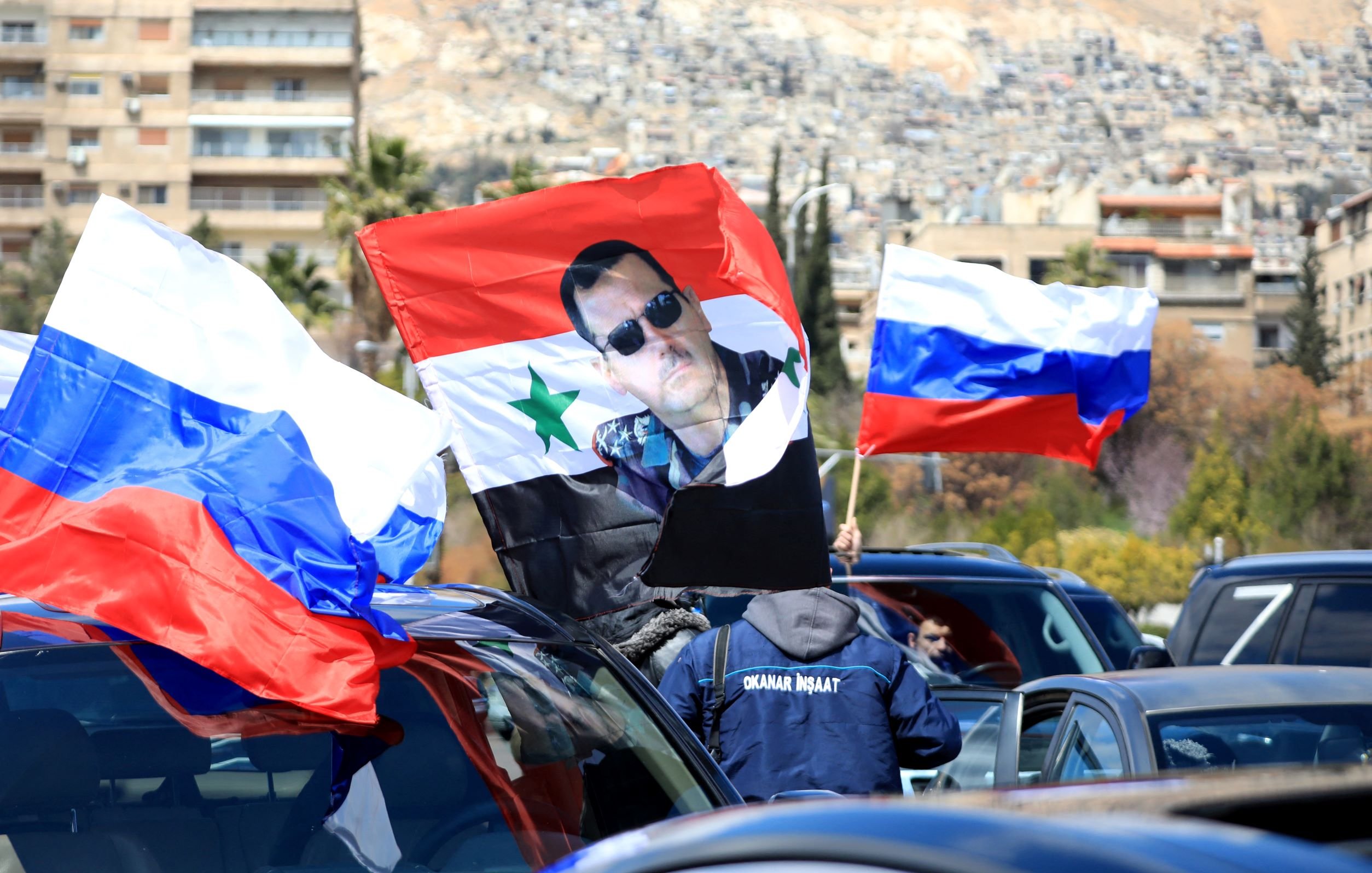 L'immagine mostra una scena di una manifestazione o di una celebrazione in cui sono presenti diverse bandiere. In primo piano si possono vedere bandiere della Russia e una bandiera con un'immagine di un individuo che indossa occhiali da sole e ha un'icona della Siria. Le auto nel contesto suggeriscono che la manifestazione potrebbe avere una certa rilevanza politica o culturale. Sullo sfondo si notano edifici e una zona collinare.
