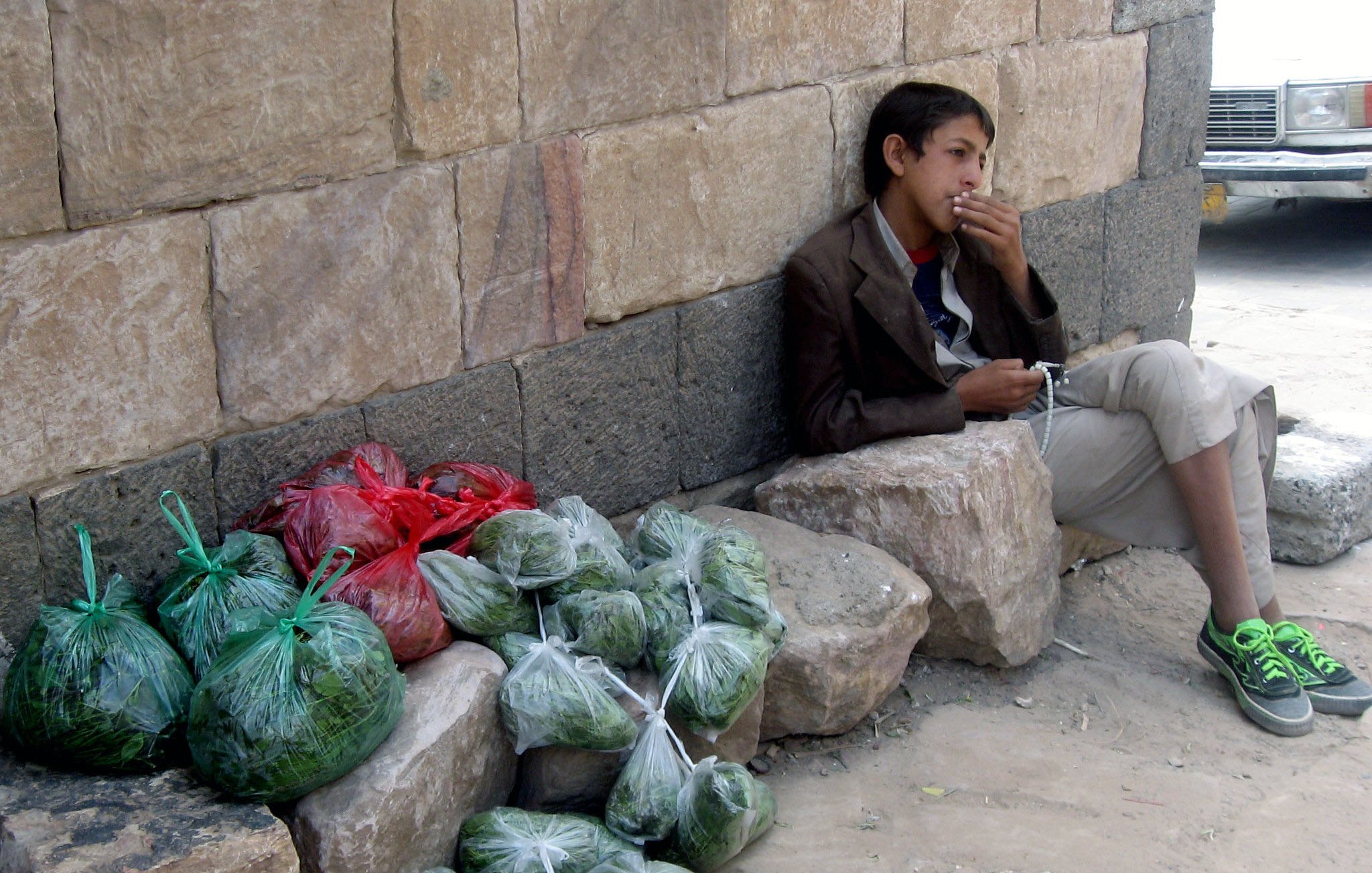 L'image montre un jeune garçon assis contre un mur en pierre. Il porte une veste et des pantalons clairs, et il semble fumer. À côté de lui, il y a plusieurs sacs transparentes contenant probablement des légumes ou des herbes, empilés sur le sol. Les sacs sont de différentes couleurs, principalement verts avec quelques rouges, et ils sont disposés sur des roches à ses côtés. L'environnement semble urbain, avec une route visible en arrière-plan.