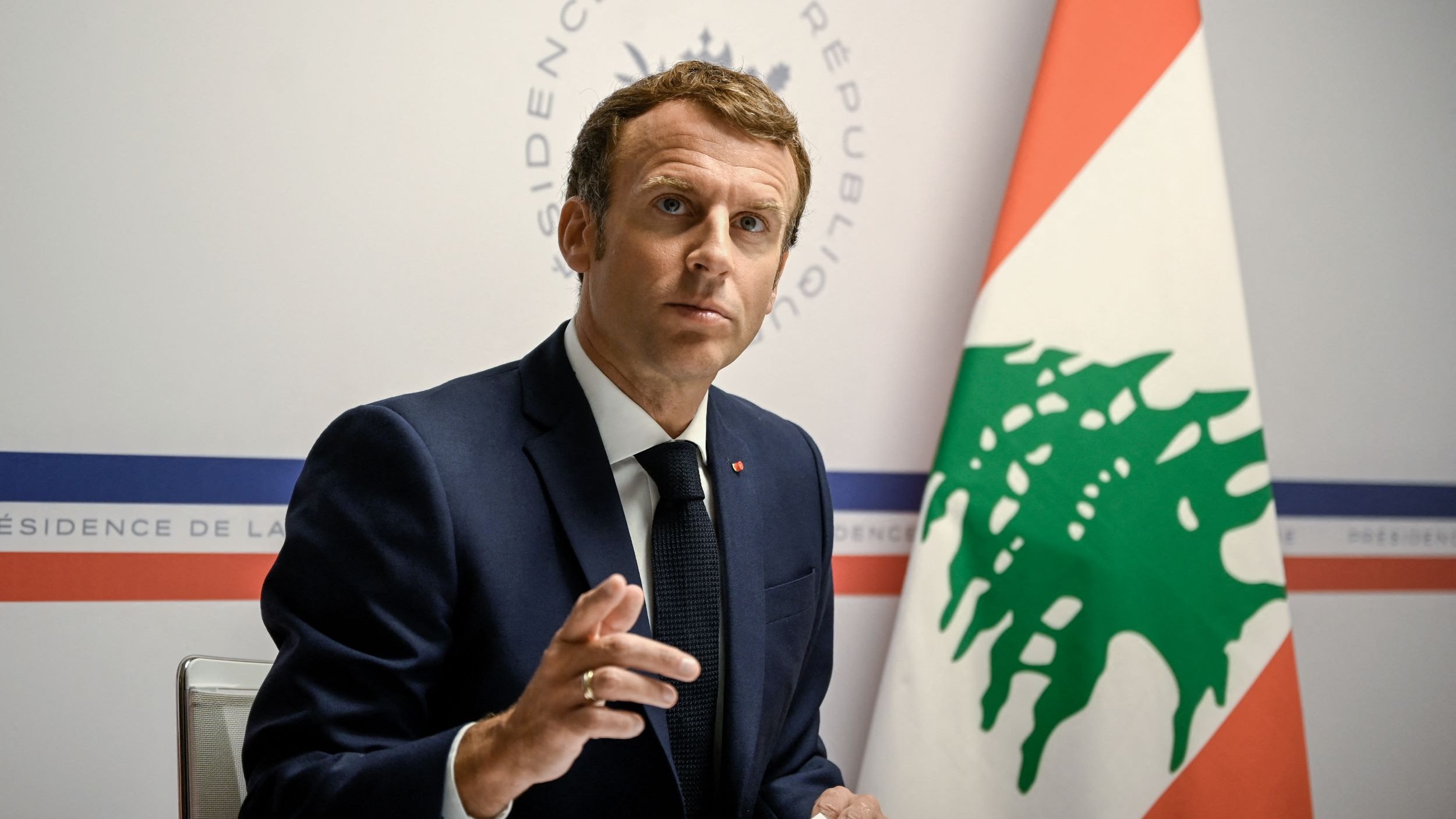 L'image montre un homme en costume, assis à une table, s'adressant à un public. Il semble être en train de faire un discours ou de répondre à des questions, avec une expression sérieuse. En arrière-plan, on peut voir un drapeau libanais ainsi qu'un logo indiquant "Présidence de la République". L'ambiance de l'image évoque un contexte officiel et diplomatique.