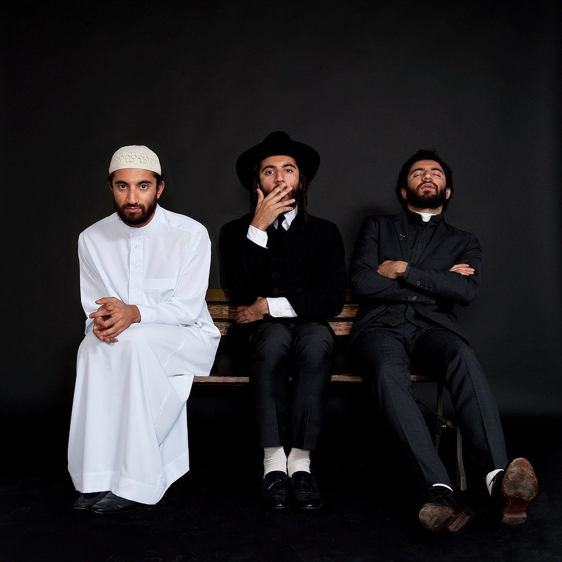L'image montre trois hommes assis sur un banc, chacun portant un style vestimentaire distinct. À gauche, un homme en blanc, portant une tenue traditionnelle et un couvre-chef. Au centre, un homme habillé de manière formelle, avec un chapeau noir et une tenue sombre. À droite, un homme également en costume, avec une expression pensive. Le fond est noir, ce qui met en valeur les vêtements et les postures des sujets.