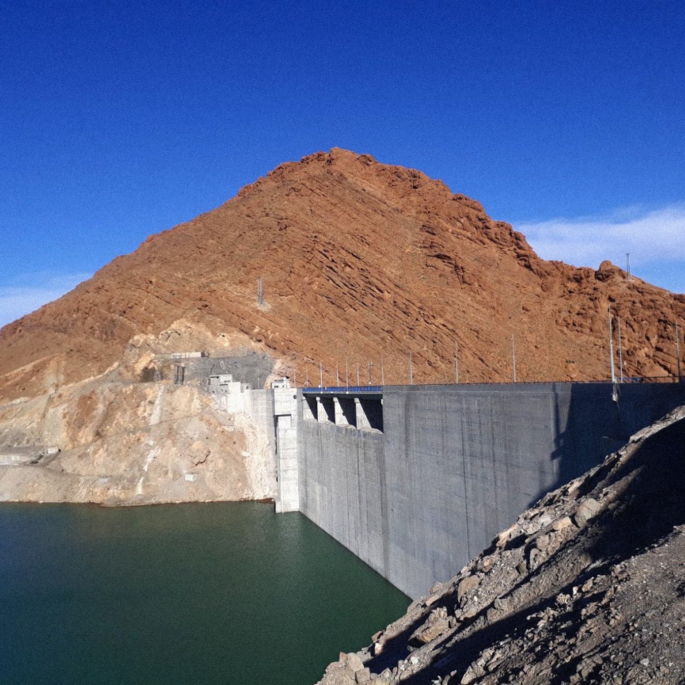 L'image montre un barrage construit sur un cours d'eau, avec une grande structure en béton qui s'étend le long de la rive. À l'arrière-plan, on voit une montagne aride, aux pentes abruptes et aux teintes brunes. Le ciel est dégagé, d'un bleu vif, et l'eau du réservoir est calme et d'une teinte verte-bleue. Le paysage semble sec et stérile, typique des régions désertiques.