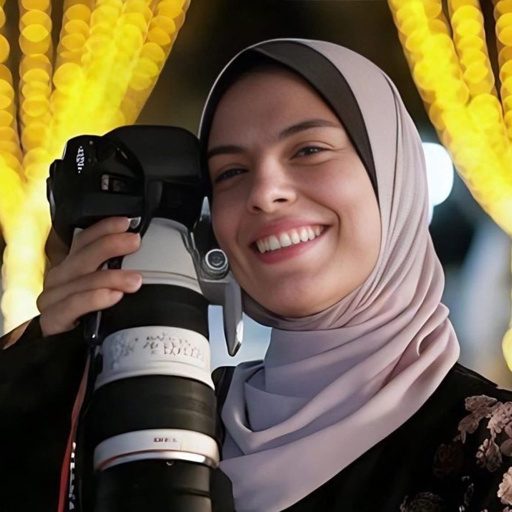 زنی با حجاب، دوربین بزرگ در دست، لبخند بر لب، در پس‌زمینه‌ای با چراغ‌های زینتی.