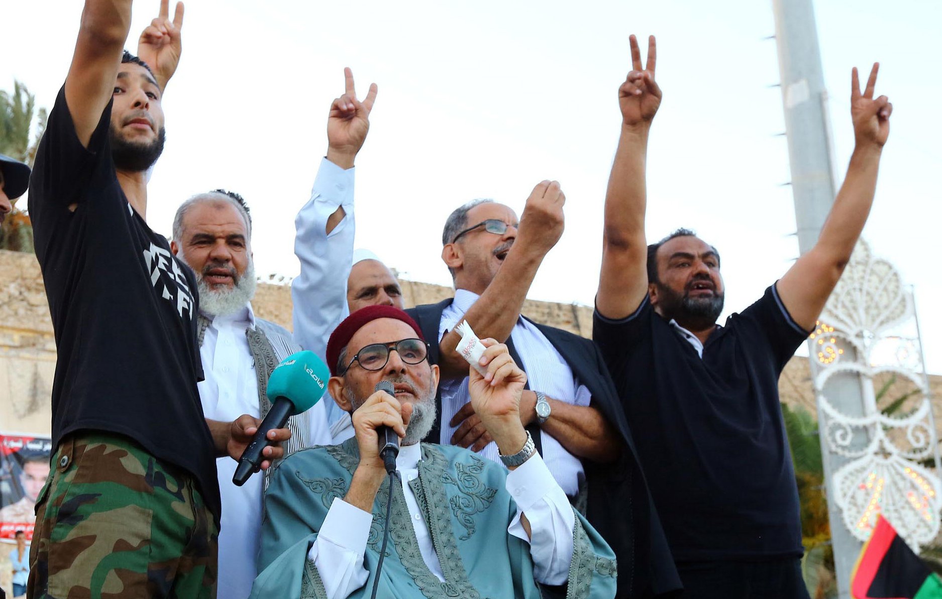 L'image montre un groupe d'hommes réunis lors d'un événement. Un homme au premier plan, portant une tenue traditionnelle, s'exprime tout en levant un symbole de paix avec ses mains. Les autres hommes en arrière-plan lèvent également les mains, probablement en signe de soutien ou de solidarité. L'ambiance semble festive et engagée, suggérant un moment de rassemblement ou de protestation. Des drapeaux ou des affiches pourraient être visibles, accentuant le contexte de l'événement.