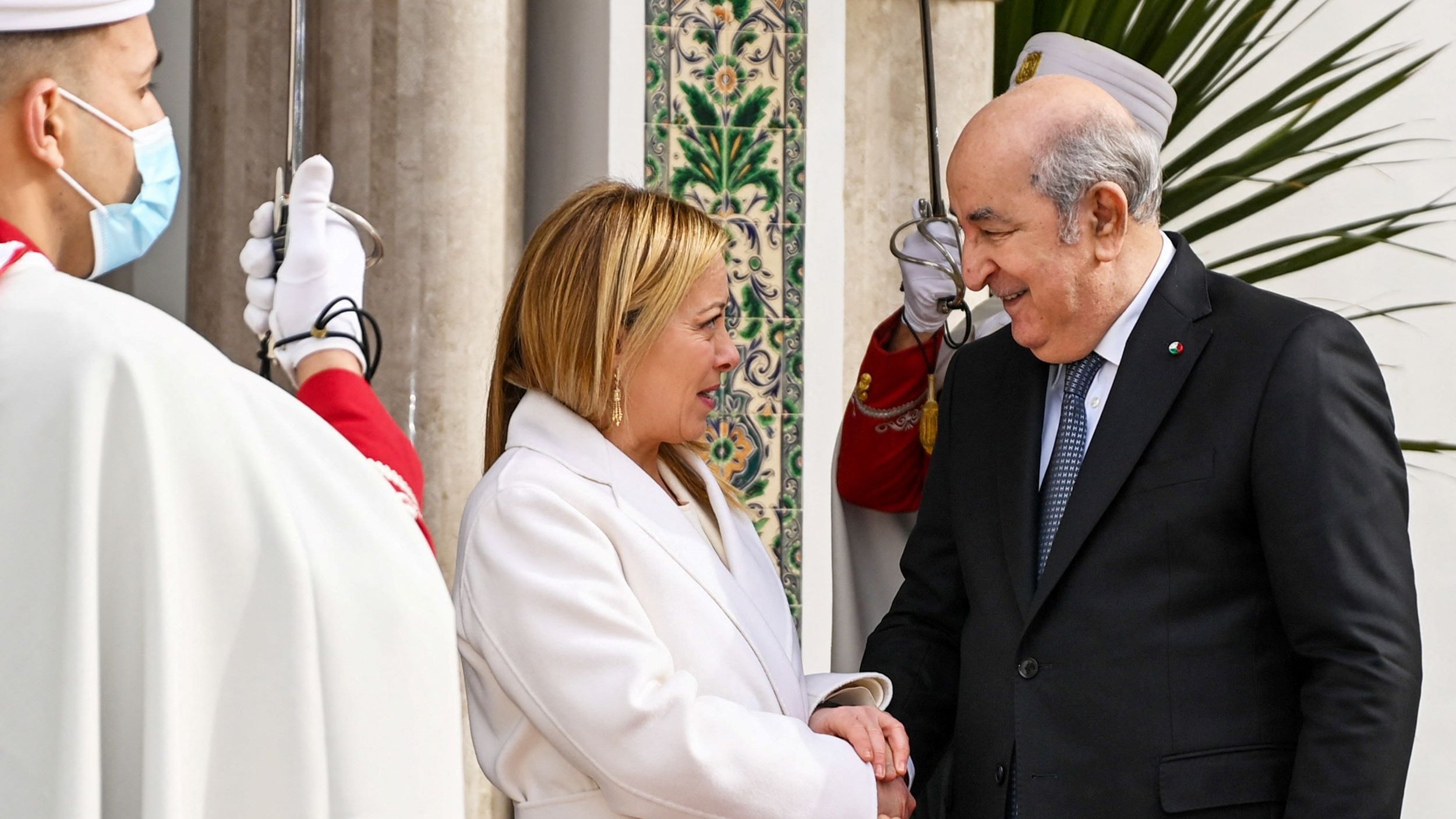L'image montre deux personnes se serrant la main avec des sourires. Une femme, habillée en blanc, semble accueillante, tandis qu'un homme en costume sombre lui répond avec un sourire. En arrière-plan, on peut voir des gardes en uniforme et des éléments d'architecture décorative, ce qui suggère un contexte officiel, probablement une rencontre diplomatique. L'atmosphère est chaleureuse et conviviale.