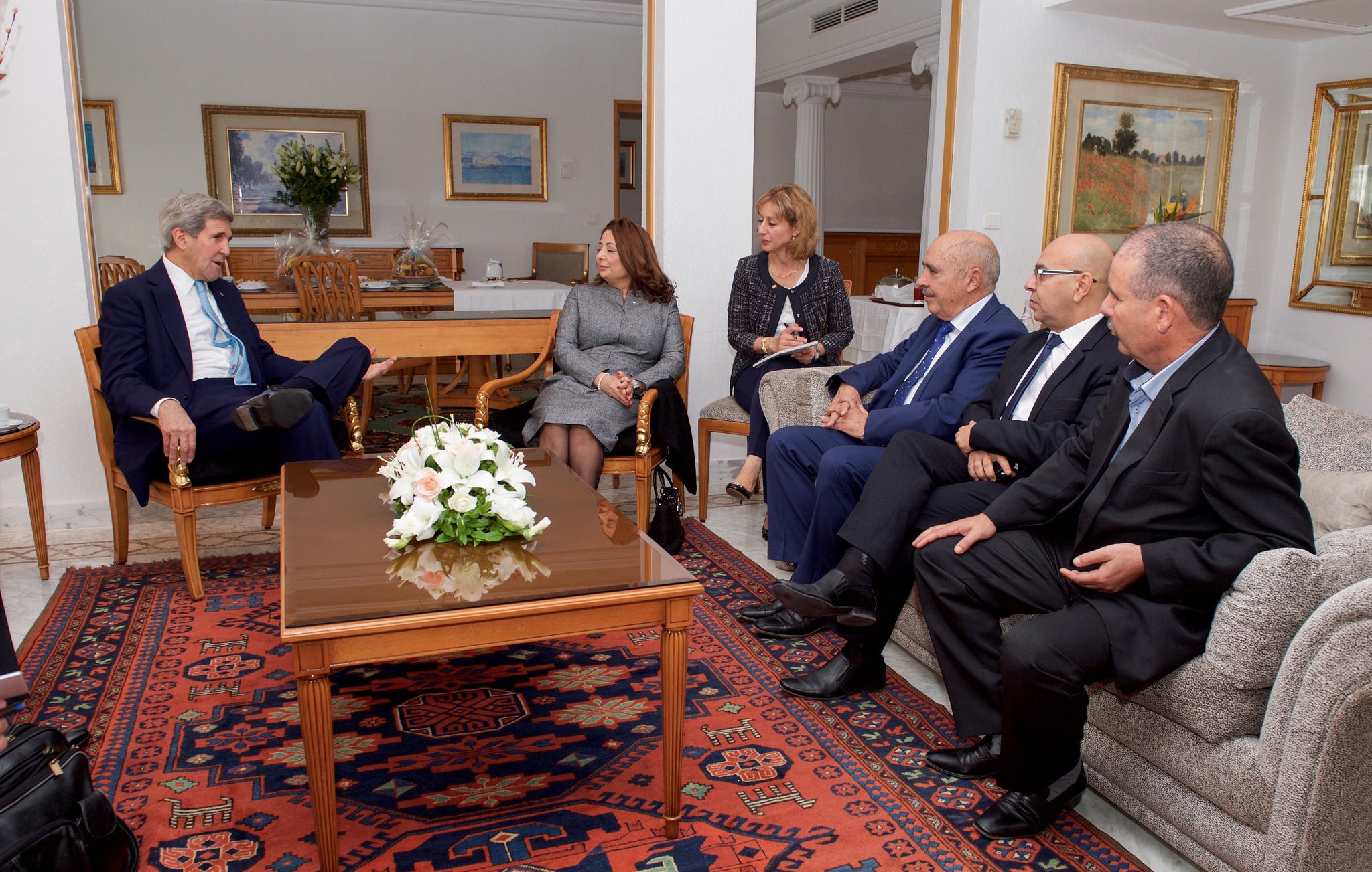 L'image montre un groupe de personnes assises dans un salon bien éclairé et décoré. Il y a une table basse au centre avec un arrangement floral blanc. À gauche, une personne est assise dans un fauteuil, écoutant attentivement. Les autres personnes sont disposées autour de la table, portant des costumes formels et engageant une conversation. Les murs sont ornés de tableaux, et le décor évoque une atmosphère professionnelle et conviviale.