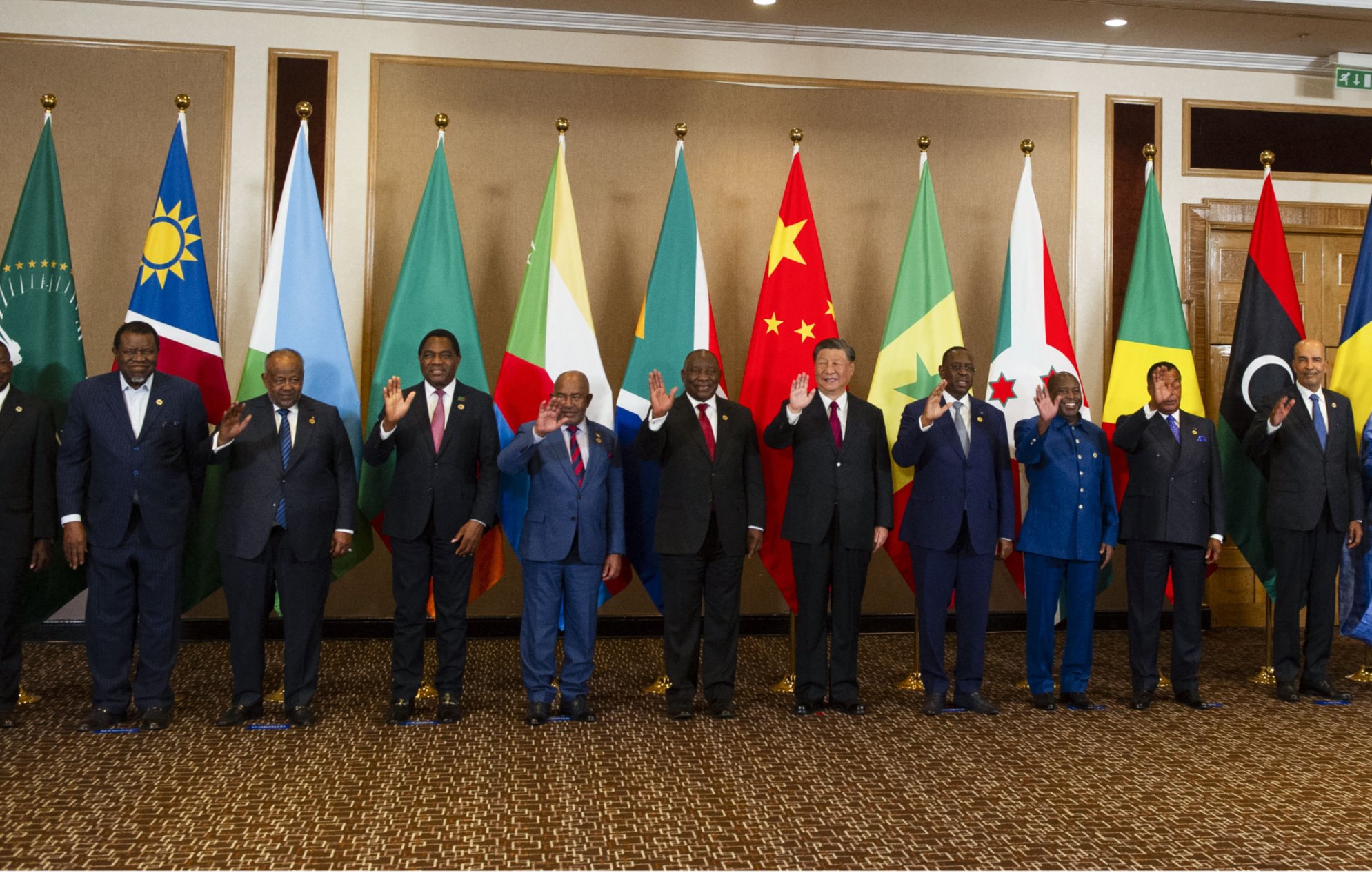 تظهر الصورة مجموعة من القادة أو الممثلين الرسميين من دول مختلفة يقفون في صف واحد. خلفهم، تُعلق أعلام تمثل الدول أو المنظمات المختلفة، بما في ذلك علم الاتحاد الأفريقي. يبدو أن الجميع يلوح بيديه في تعبير عن التحية. يرتدي القادة بدلات رسمية، وبعضهم يرتدي الملابس التقليدية. تعكس الصورة مناسبة رسمية أو قمة تجمع بين الدول.