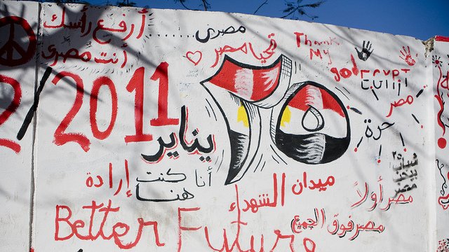 L'image montre un mur recouvert de graffitis en arabe et en anglais. Parmi les inscriptions, on trouve des références à l'Égypte, à la révolution de 2011, et des slogans tels que "Meilleur avenir" et des phrases évoquant la détermination et l'espoir. Les couleurs rouge, noir et jaune du drapeau égyptien sont visibles, renforçant le message patriotique et engagé. L'ensemble évoque une période de changement et de revendications sociales.