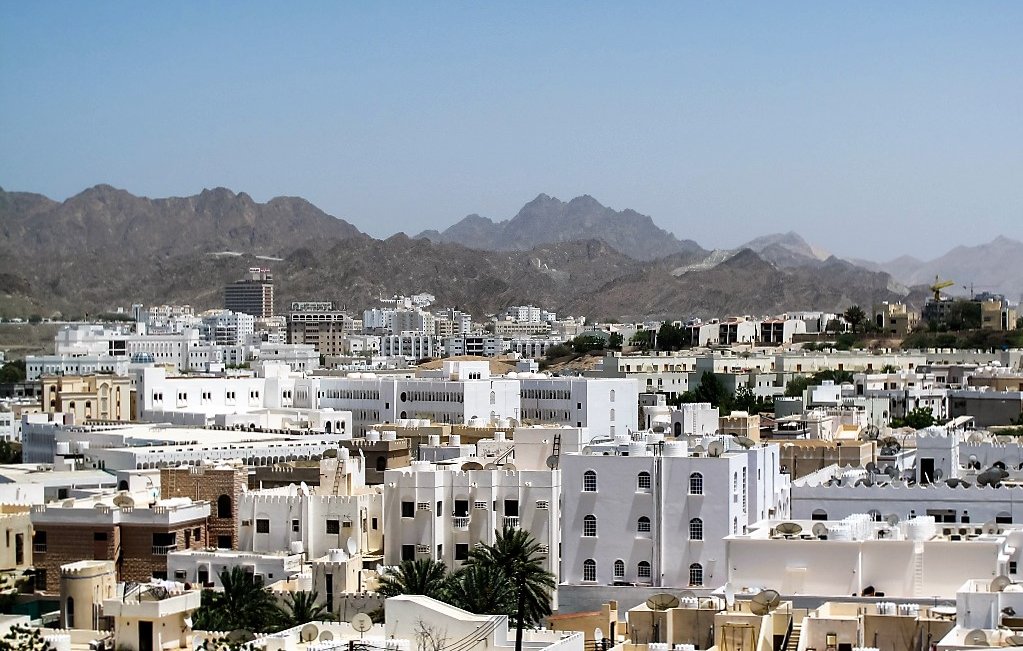 L'image montre une vue panoramique d'une ville située dans une région montagneuse. On peut voir de nombreux bâtiments aux façades blanches et crème, qui typiquement se distinguent dans un environnement aride. Les montagnes en arrière-plan ajoutent une dimension naturelle impressionnante au paysage urbain. Le ciel est dégagé et ensoleillé, accentuant la lumière qui se reflète sur les bâtiments. Cette scène évoque une atmosphère calme et sereine.
