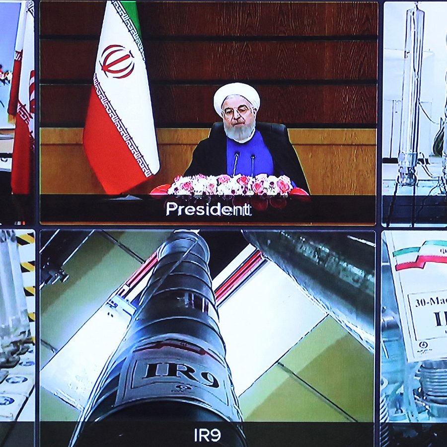 L'image montre une composition de plusieurs écrans de télévision. Au centre, on voit un homme, probablement un président, assis devant un bureau avec des fleurs, entouré de drapeaux iraniens. Sur les autres écrans, il y a des images d'installations ou d'équipements liés à la technologie nucléaire, avec des tubes et des étiquettes indiquant différentes séries de centrifugeuses nucléaires. Il y a aussi des symboles et des drapeaux qui décrivent les différentes installations mentionnées. L'ensemble semble se rapporter à un discours ou une présentation sur le programme nucléaire de l'Iran.