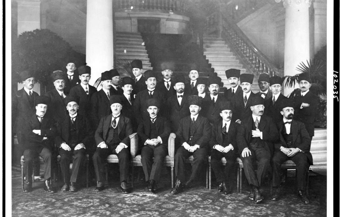 L'image présente un groupe d'hommes, probablement d'importance politique ou officielle, assis et debout dans un cadre formel. Ils portent des costumes sombres et des chapeaux traditionnels, et certains ont des moustaches. La scène se déroule dans un intérieur élégant, avec des colonnes décoratives et des plantes en pot en arrière-plan. L'atmosphère dégage une impression de solennité et de cérémonie, suggérant un événement ou une réunion importante.