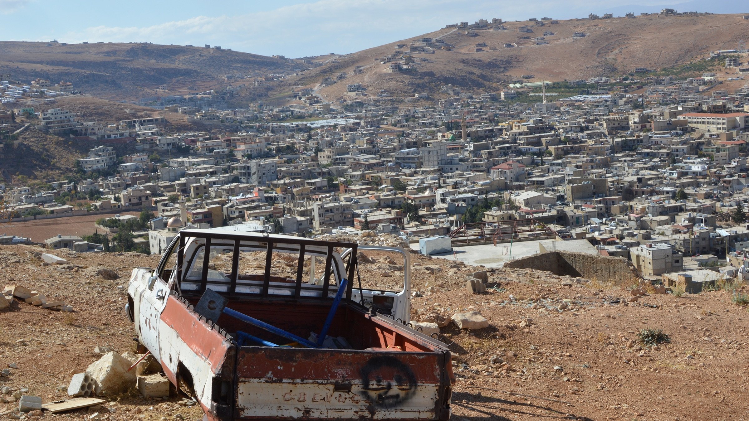 L'image montre une scène de paysage où un ancien véhicule, probablement une camionnette, est abandonné sur une colline. La camionnette est en mauvais état, avec une partie de la carrosserie rouillée et peinte en blanc et rouge. En arrière-plan, on peut voir un village ou une ville avec de nombreuses maisons, s'étendant sur les collines. Le ciel est partiellement nuageux, et le paysage environnant est aride, typique des terrains montagneux.