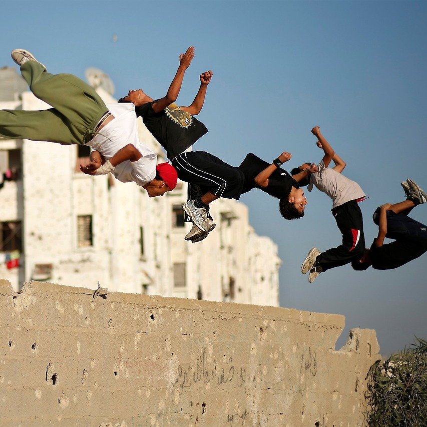 L'image montre un groupe de jeunes en train de sauter par-dessus un mur. Ils semblent s'amuser à réaliser des acrobaties, témoignant d'une grande énergie et d'une certaine audace. En arrière-plan, on aperçoit des bâtiments avec des murs marqués, ce qui donne une idée de l'environnement urbain. Le ciel est clair, ce qui contraste avec les structures des bâtiments. L'ambiance générale de l'image est dynamique et vivante, capturant un moment de loisirs et de camaraderie entre les adolescents.