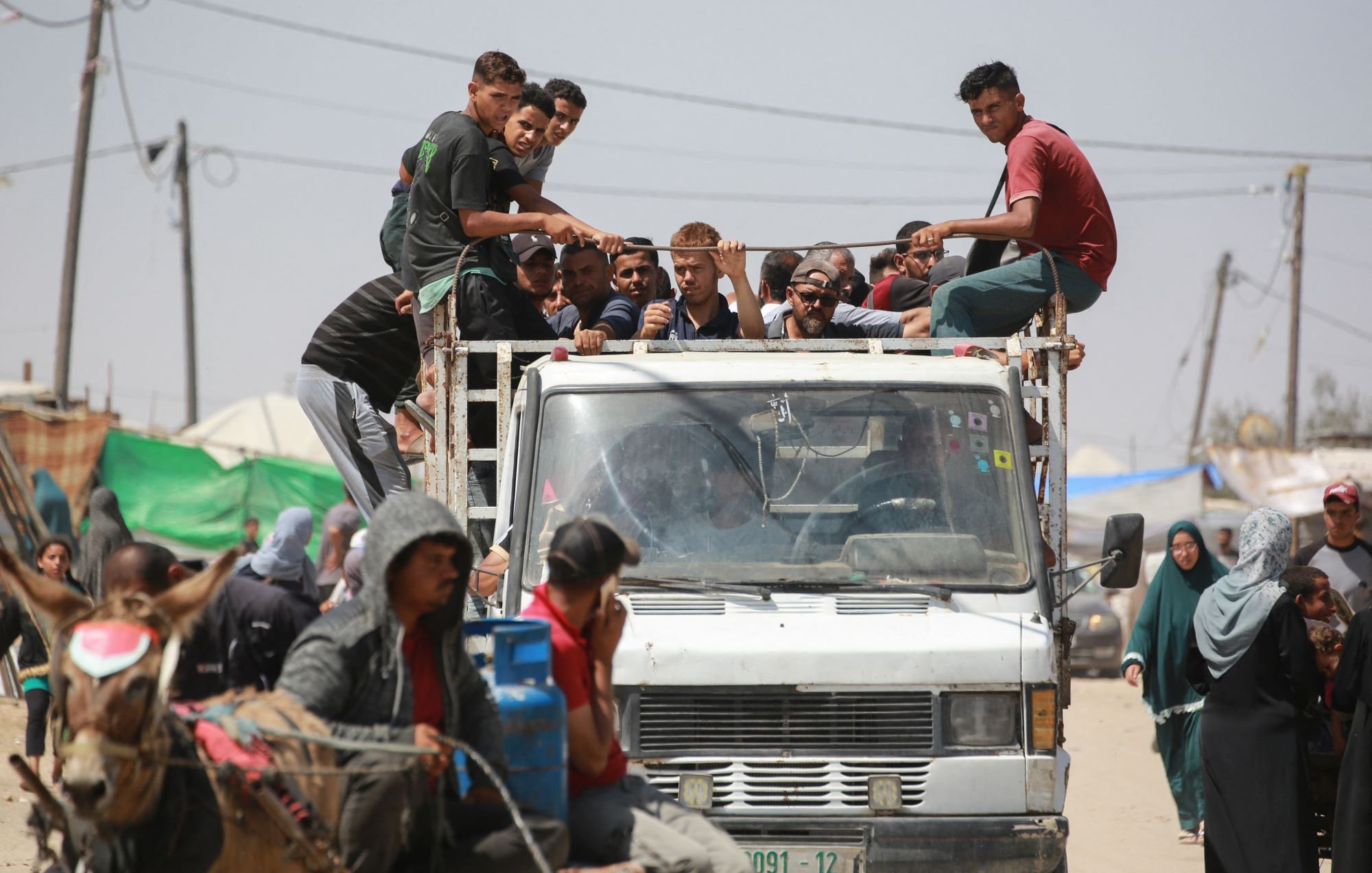 در این تصویر، گروهی از جوانان در حال نشستن بر روی یک کامیون هستند. برخی از آن‌ها در قسمت بار کامیون نشسته‌اند، در حالی که دیگران به طرف خودرویی که در حرکت است، نگاه می‌کنند. در جلوی تصویر، چند نفر در کنار کامیون و همچنین زنانی با پوشش‌های سنتی مشاهده می‌شوند. در پس‌زمینه، سایه‌هایی از چادرها و همچنین سیم‌کشی‌های برق دیده می‌شود که نشان‌دهنده یک منطقه پرجمعیت و شاید غیررسمی است. احساسی از شلوغی و جنب و جوش از صحنه به‌طور کلی مشهود است.