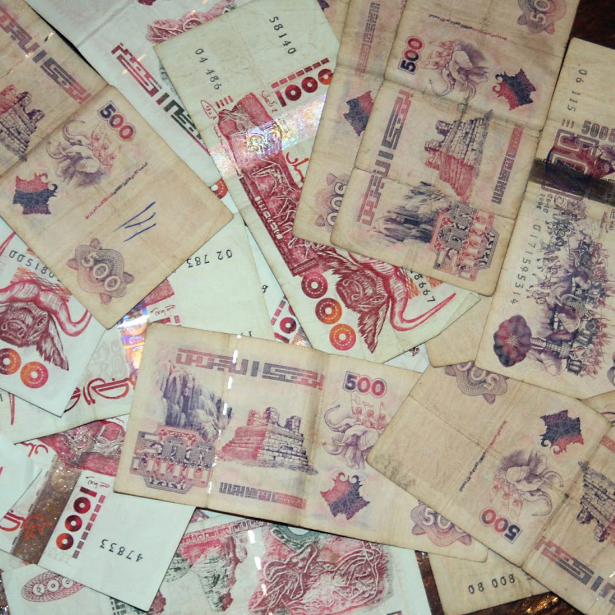 L'image montre un tas de billets de banque éparpillés, principalement de couleur beige avec des motifs et des textures variés. Certains billets sont de 500 unités, affichant des illustrations ou des inscriptions.