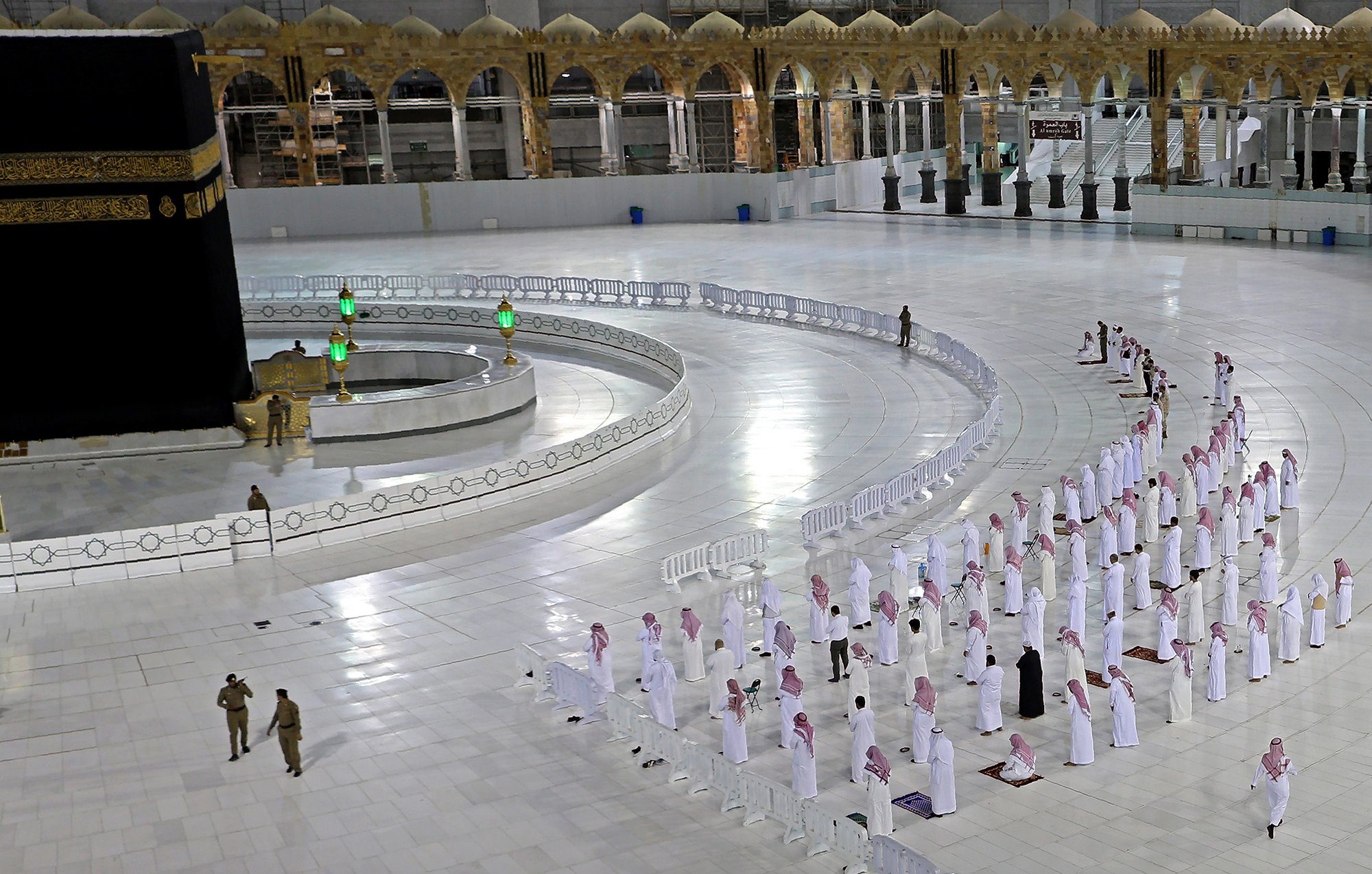 L'image montre une scène à la Kaaba, située à La Mecque. Plusieurs personnes en vêtements traditionnels blancs, souvent appelés " Ihram", sont disposées en rangées, apparemment en train de prier. On peut voir la Kaaba, un cube noir avec des détails dorés, au centre du sanctuaire. Des barrières de sécurité sont présentes pour délimiter les zones, et l'espace est vaste et lumineux, avec des éléments architecturaux en arrière-plan. Cette image évoque un moment de recueillement et de spiritualité dans un lieu sacré pour les musulmans.