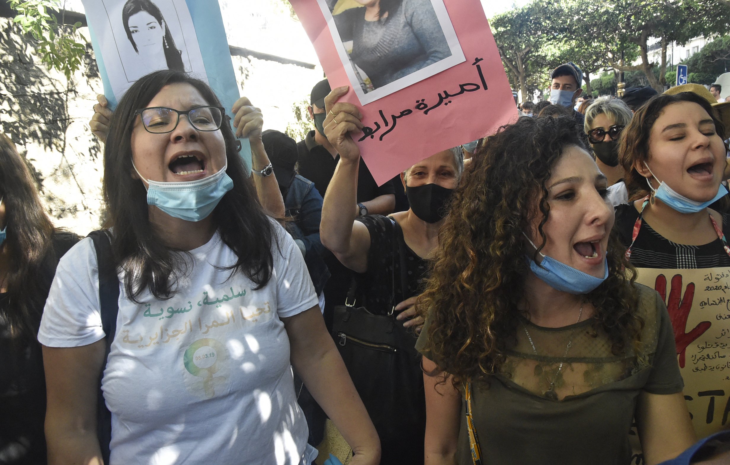 L'immagine mostra un gruppo di persone che partecipa a una manifestazione. Alcuni manifestanti tengono in mano cartelli con fotografie e slogan, mentre gridano in segno di protesta. Le persone indossano mascherine e sembrano essere molto coinvolte emotivamente, esprimendo le loro richieste con forza. Il contesto sembra essere urbano, con alberi e edifici sullo sfondo.