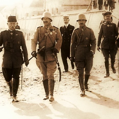 L'image montre un groupe de militaires marchant ensemble sur un chemin, probablement dans un contexte historique. Les hommes portent des uniformes militaires traditionnels, avec des médailles visibles sur certains d'entre eux. Ils semblent avoir un air sérieux et solennel. En arrière-plan, on distingue des tentes, ce qui pourrait indiquer un camp ou une retraite militaire. L'atmosphère de la scène est marquée par la formalité et l'importance de l'événement.