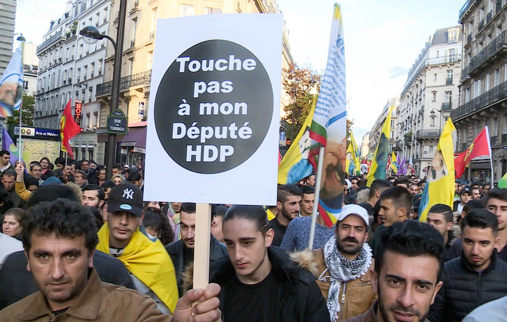 L'image montre une manifestation de foule dans une rue, où des personnes tiennent des pancartes et des drapeaux. Au premier plan, un manifestant porte une grande pancarte avec le message "Touche pas à mon Député HDP". L'ambiance semble être celle d'une protestation politique, impliquant probablement des questions liées au parti HDP (Parti démocratique des peuples) en Turquie. Les participants semblent engagés et déterminés.
