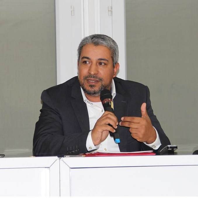 L'image montre un homme assis à une table, tenant un microphone. Il semble engager une discussion ou faire une présentation. Il porte un costume sombre avec une chemise claire et a des cheveux grisonnants. L'environnement autour de lui semble être celui d'une conférence ou d'un événement formel, avec une lumière claire et des fenêtres en arrière-plan.