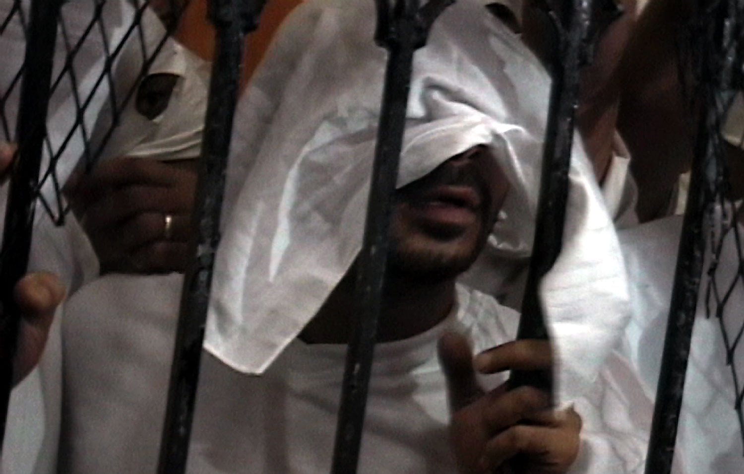 L'image montre une personne avec un regard partiellement caché par un tissu blanc. Elle se trouve derrière des barreaux, ce qui suggère une situation de confinement ou de détention. Les mains de la personne sont visibles, tenant les barreaux, et le fond de l'image semble flou, mettant l'accent sur la figure centrale. L'atmosphère de l'image peut évoquer des sentiments d'isolement ou de détresse.