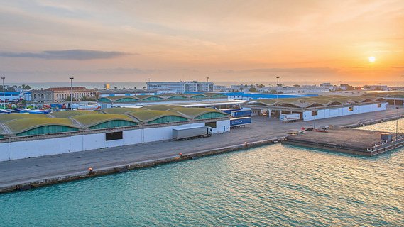 L'image montre un port lors d'un coucher de soleil, avec un paysage industriel. On peut voir des entrepôts aux toits arrondis, bordés par l'eau. Des véhicules et des conteneurs sont visibles, indiquant une activité portuaire. Le ciel est teinté de couleurs chaudes, créant une atmosphère paisible et accueillante.
