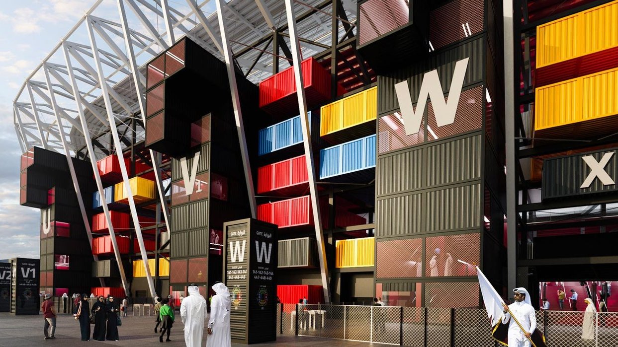 الصورة تظهر تصميمًا عصريًا لمنطقة بجانب ملعب أو منشأة رياضية. يتميز التصميم بألوان زاهية تتضمن الصناديق الملونة بالأسود والأحمر والأصفر والأزرق. هناك أيضًا مدخل واسع مع شعارات واضحة. في الجزء الأمامي، يظهر بعض الأشخاص يرتدون الملابس التقليدية، مما يشير إلى إمكانية وقوع الحدث في منطقة عربية أو إسلامية. السماء تتسم بصبغة رمادية توحي بوقت الغسق.