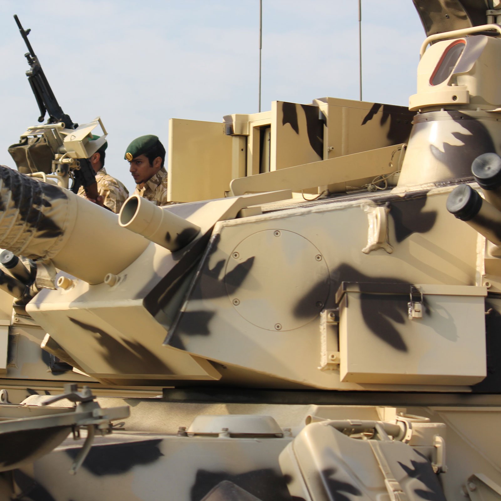 L'image montre un char militaire avec un camouflage dans des tons de beige et de marron. On peut voir un soldat à proximité, portant un uniforme militaire et un casque. Le char est équipé d'un canon en position, et il semble être prêt pour une opération. L'environnement semble être en plein air, peut-être lors d'exercices ou d'une parade militaire, avec un ciel clair en arrière-plan.