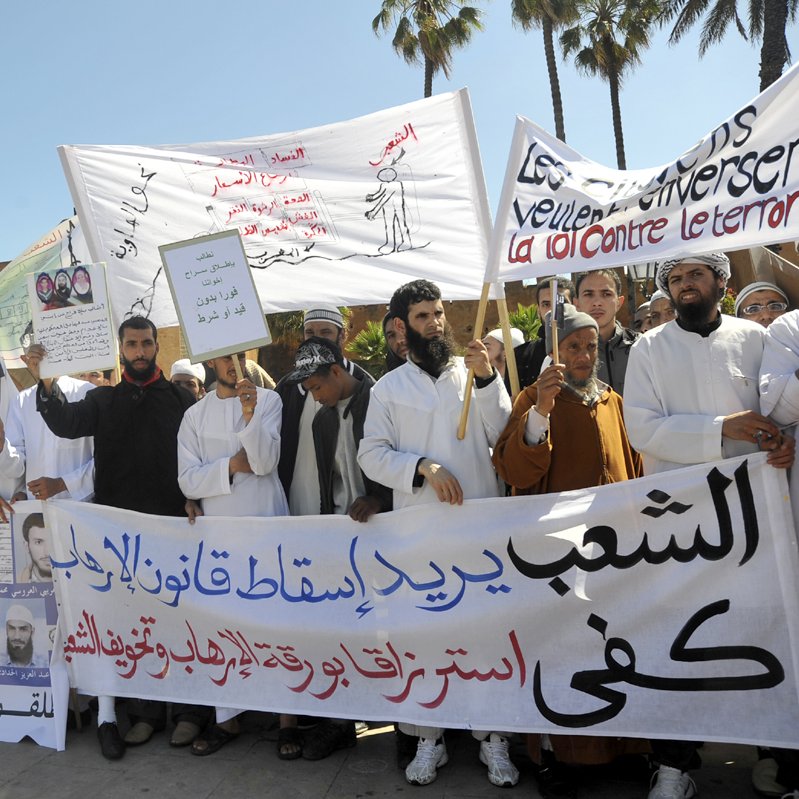 L'image montre une manifestation avec un groupe de personnes tenant des pancartes et des banderoles. Les manifestants semblent exprimer des revendications politiques ou sociales, avec des slogans écrits en arabe et en français. On peut voir des visages déterminés et des gestes de protestation. La scène est entourée d'arbres, suggérant un environnement urbain. L'atmosphère est chargée d'émotion, reflétant l'engagement des participants.