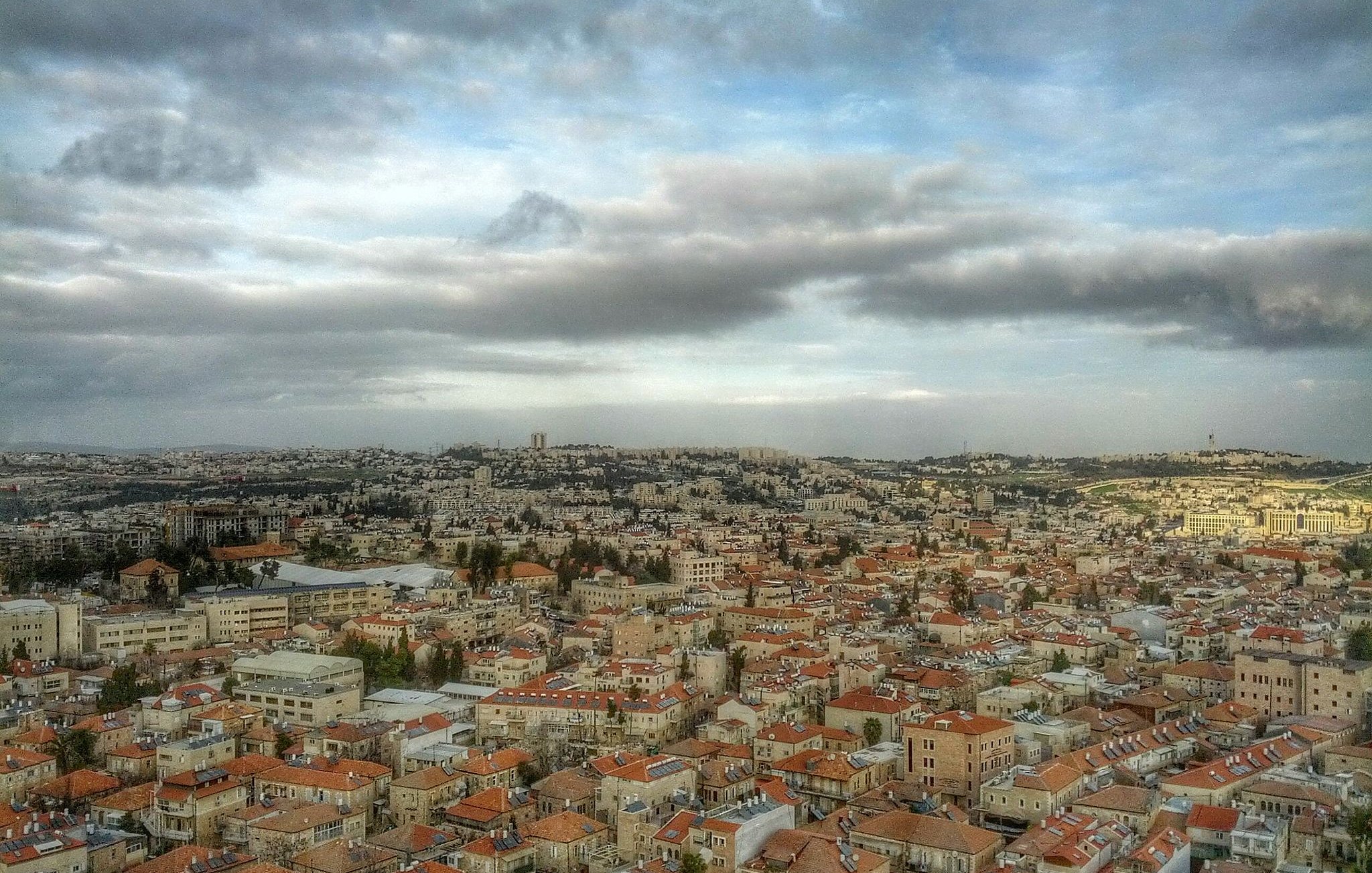 L'image montre une vue panoramique d'une ville, probablement Jérusalem, avec des bâtiments disposés sur plusieurs collines. Les toits sont principalement en tuiles rouges, et il y a de nombreux édifices en pierre. Le ciel est nuageux, ajoutant à l'ambiance de la scène, avec une lumière douce qui éclaire la ville. On peut également apercevoir des zones de verdure entre les bâtiments, ce qui ajoute de la profondeur au paysage urbain.