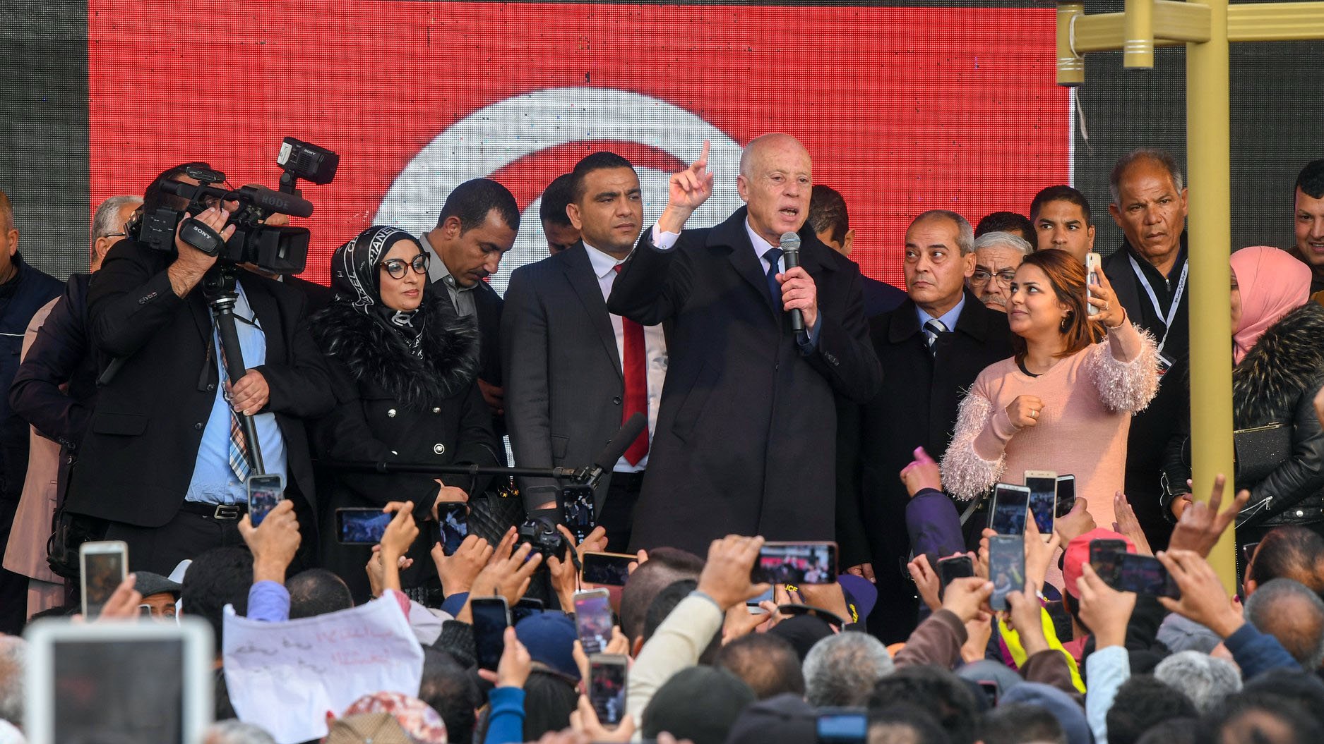 تظهر في الصورة حشود كبيرة من الناس تجمعوا في مكان عام. في المقدمة، يظهر شخص يقف على منصة ويتحدث، بينما يمسك العديد من الحضور هواتفهم المحمولة لتوثيق اللحظة. يحيط به مجموعة من الأشخاص، وبعضهم يبدو مشغولاً بالتقاط الصور أو الفيديو. خلف المنصة، توجد لافتة تحمل الألوان الوطنية. الأجواء توحي بالحماس والاهتمام من الحضور.