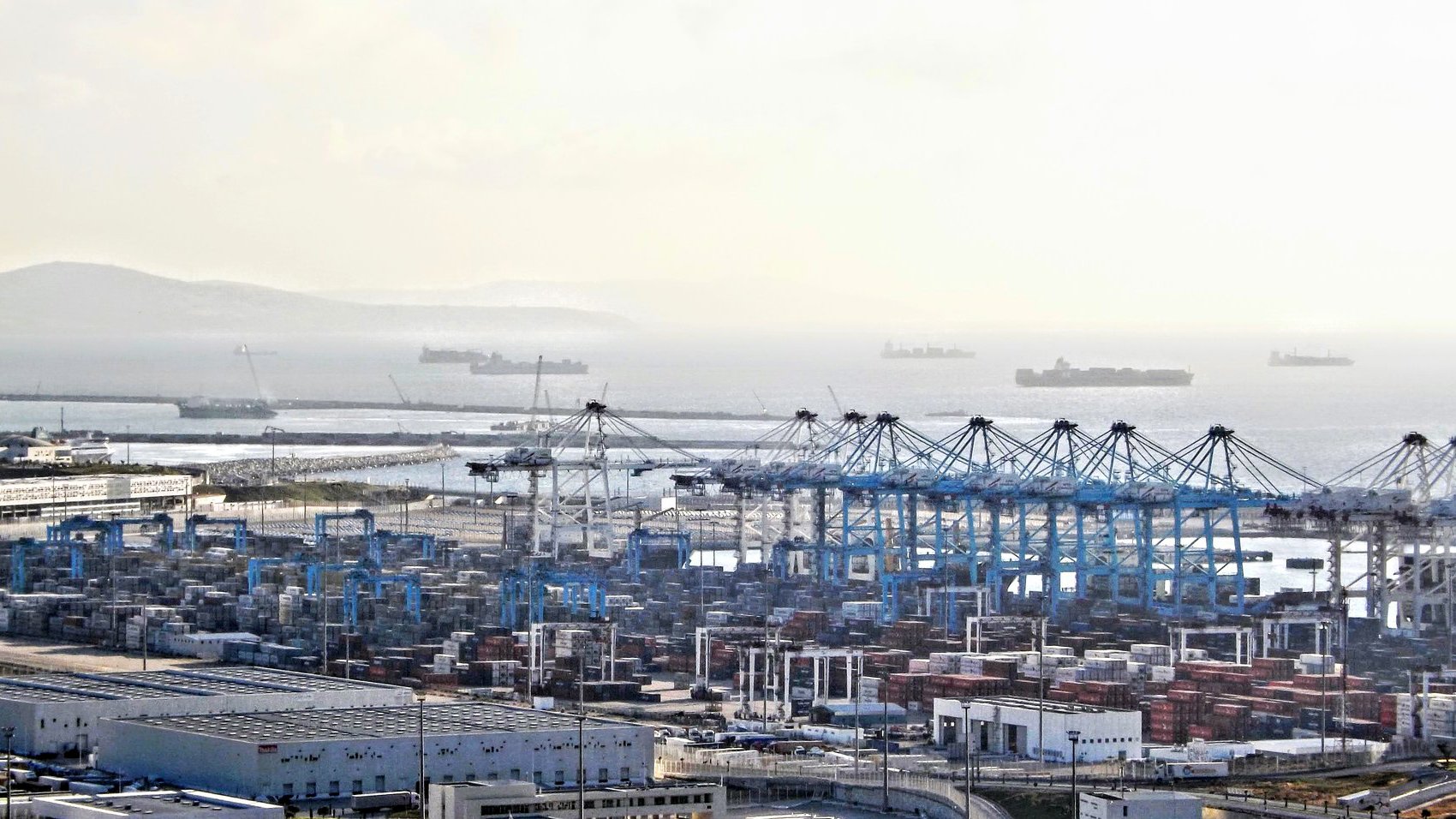 L'image montre un port maritime avec des conteneurs empilés, des grues de chargement et plusieurs navires au large. On peut voir des bâtiments et des installations portuaires. L'horizon est légèrement brumeux, créant une atmosphère calme, et des bateaux de différentes tailles sont visibles sur la mer. L'ensemble dépeint une scène de transport maritime et de logistique.
