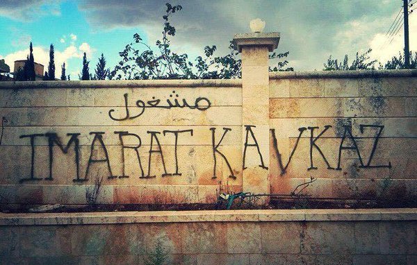 L'image montre un mur en pierres, probablement d'un bâtiment ou d'une clôture. Sur ce mur, il y a des graffitis qui sont en arabe et en lettres latines. Le mot en arabe semble se traduire par "travail" ou "emploi", tandis que "IMARAT KAVKAZ" est écrit en grandes lettres au centre. L'environnement autour du mur est nature, avec quelques arbres et un ciel nuageux, ce qui donne une ambiance un peu mélancolique ou chargée.