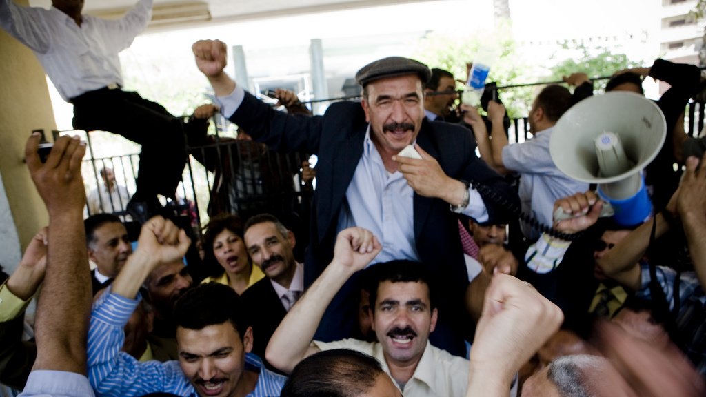 L'image montre une grande foule de personnes rassemblées, manifestant avec enthousiasme. Au centre, un homme souriant est porté sur les épaules d'un autre, levant le poing en signe de victoire. Les autres participants expriment également leur excitation, certains levant les bras et criant. La scène semble vibrante et dynamique, indiquant un moment de célébration ou de rassemblement collectif. Des panneaux et un mégaphone sont également visibles, suggérant une forme de discours ou de protestation.