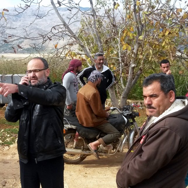 L'image montre un groupe d'hommes dans un environnement rural, probablement en plein air avec des arbres autour. Un homme, habillé avec une veste noire, est au centre, en train de parler au téléphone et de pointer du doigt dans une direction. D'autres hommes sont dispersés autour de lui, certains se tenant debout, d'autres assis sur une moto. L'arrière-plan montre des collines ou des montagnes, suggérant une zone naturelle. L'atmosphère semble détendue mais attentive, comme si les hommes discutaient d'un sujet important.