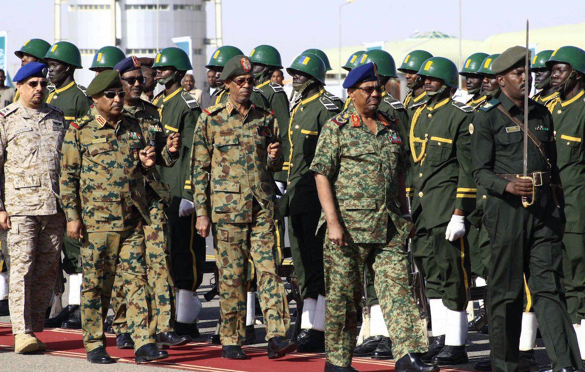 L'image montre un groupe de militaires en uniforme, marchant en formation. Les soldats portent des uniformes variés, notamment des uniformes camouflés et d'autres de couleur kaki, ornés de médailles et d'insignes. Certains militaires portent des casques ou des bérets. Au premier plan, des officiers semblent inspecter les troupes, tandis qu'une formation de soldats en arrière-plan est prête, alignée sur une surface rouge, probablement lors d'une cérémonie officielle. L'ambiance semble solennelle et rituelle, caractéristique d'une parade militaire.