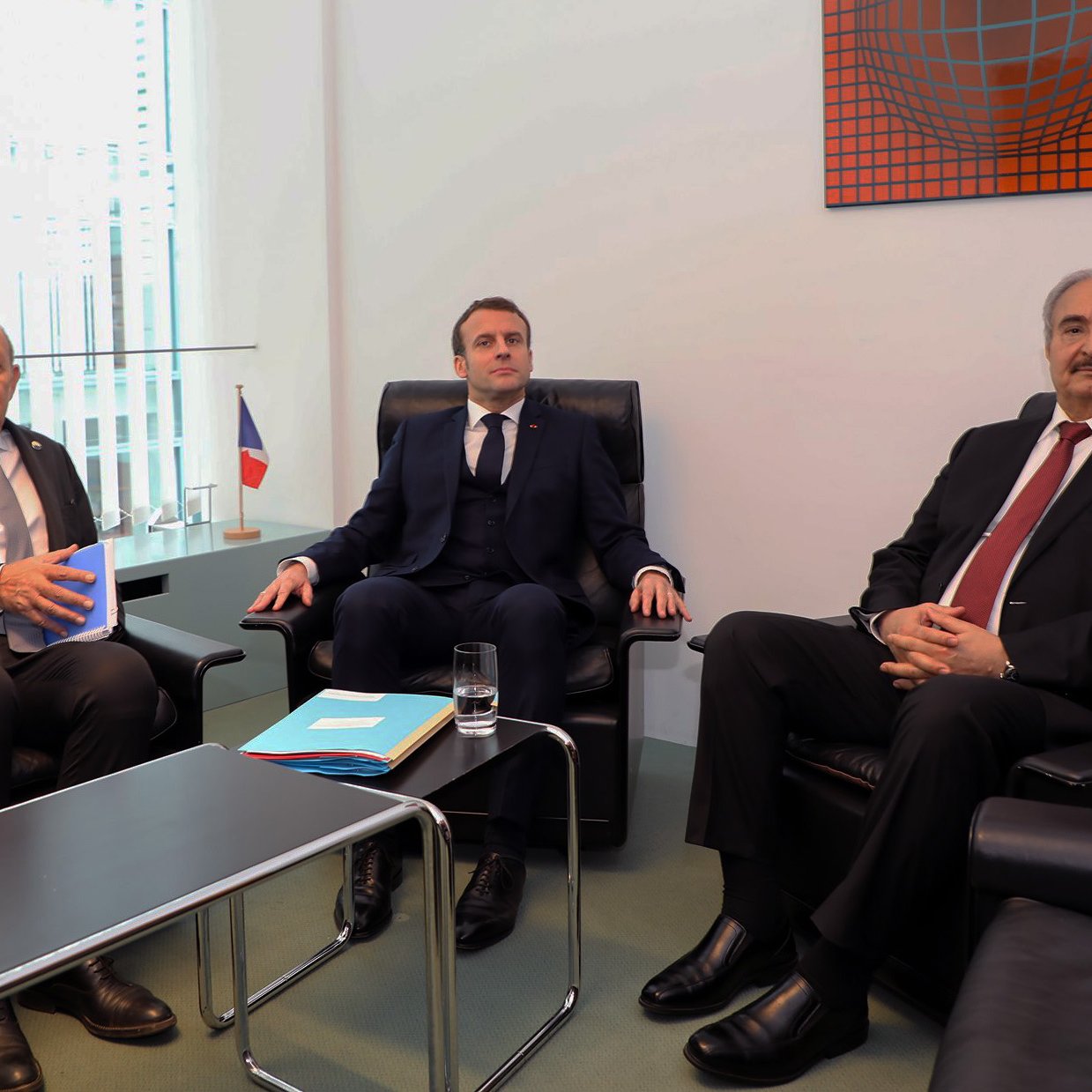 در این تصویر سه نفر نشسته‌اند. یکی از آنها در سمت چپ یک مرد با موهای خاکستری و در حال صحبت است. در وسط، مردی با کت و کراوات تیره نشسته که به نظر می‌رسد به موضوعی توجه می‌کند. شخص سوم در سمت راست یک مرد با موهای سیاه و چهره‌ای جدی است که او نیز در حال حاضر در نشستی قرار دارد. پس‌زمینه تصویر شامل پنجره‌هایی است که نور طبیعی را به اتاق وارد می‌کند. در کنار این افراد، پرچمی از فرانسه به چشم می‌خورد.