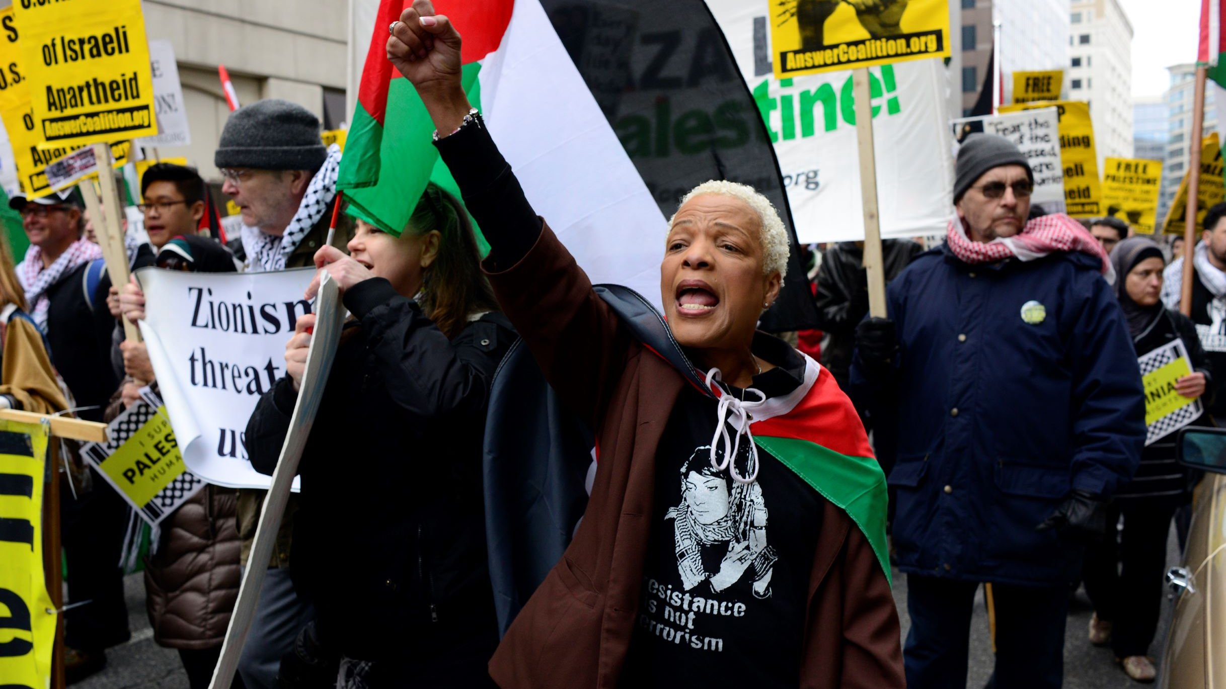 L'image montre une manifestation avec un groupe de personnes brandissant des pancartes et des drapeaux. Une femme au premier plan, portant un t-shirt avec une inscription, lève le poing en criant des slogans. Les manifestants semblent exprimer leur soutien à la cause palestinienne. On peut apercevoir des drapeaux aux couleurs palestiniennes, ainsi que des affiches dénonçant le sionisme et appelant à la résistance. L'ambiance semble dynamique et engagée.