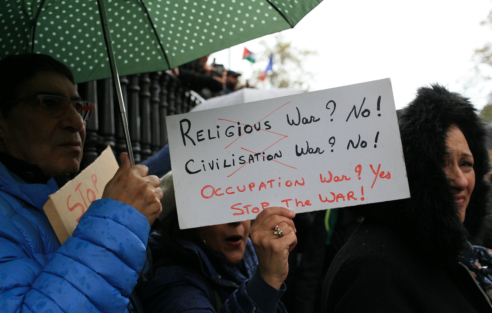 في الصورة، يظهر مجموعة من الأشخاص في تظاهرة، حيث يحمل أحدهم لافتة مكتوب عليها "الحرب الدينية؟ لا! الحرب الحضارية؟ لا! حرب الاحتلال؟ نعم! اوقفوا الحرب!"، بينما يحمل آخر مظلة خضراء. التعبيرات على وجوه المتظاهرين تدل على الإصرار والمشاركة الفعالة في الاحتجاج. الأجواء تتسم بالجدية مع خلفية غائمة أو ممطرة.