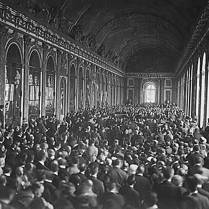 L'image montre une vaste salle intérieure, probablement un palais, remplie d'un grand nombre de personnes. On peut voir des rangées de chaises disposées, et les murs sont ornés de grandes fenêtres et de détails architecturaux. La foule semble rassemblée pour un événement ou une cérémonie importante, créant une atmosphère dense et dynamique dans cet espace majestueux. La lumière pénètre par les fenêtres, ajoutant une ambiance solennelle à la scène.