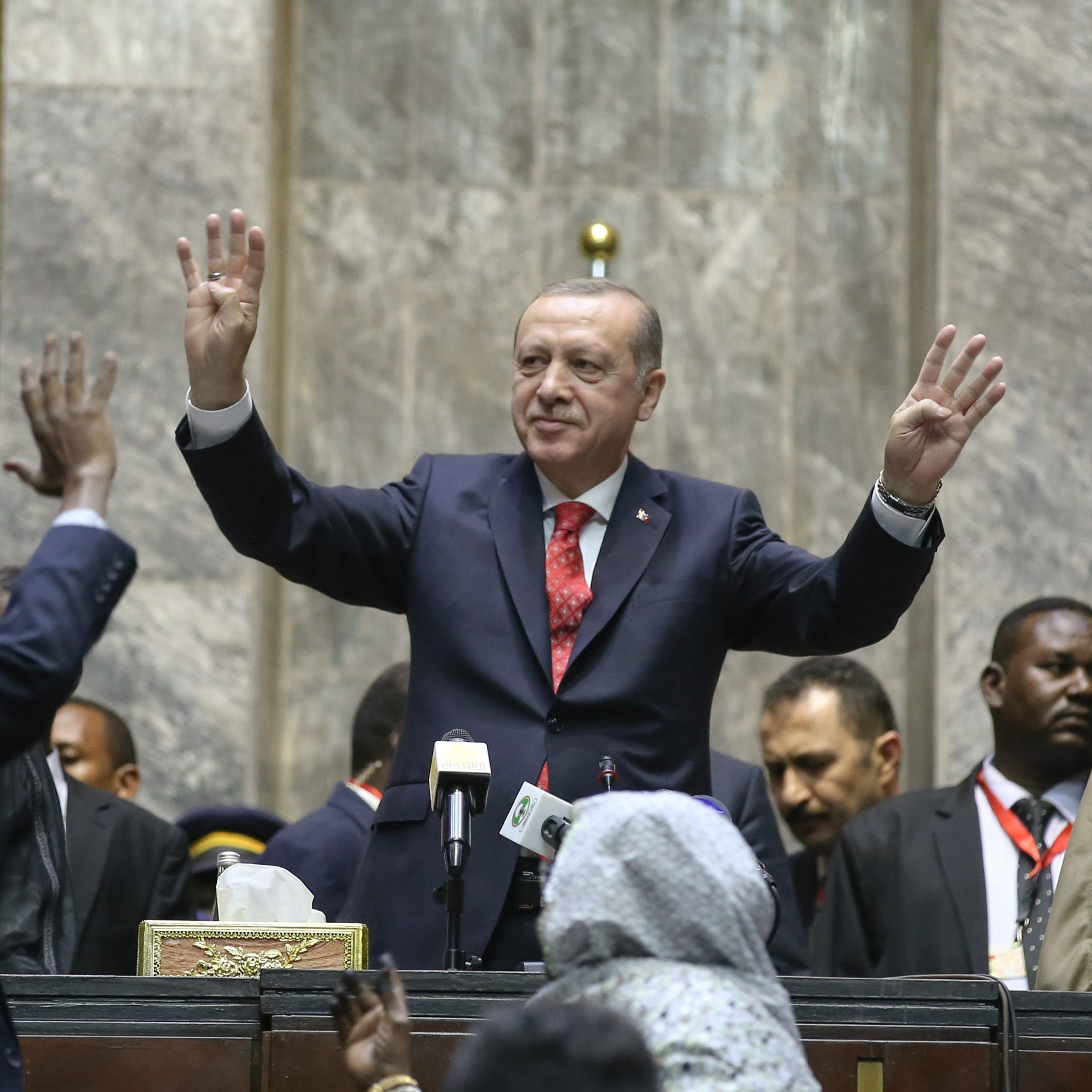 L'image montre un homme debout au centre, levant les mains en signe de salutation ou de triomphe. Il est entouré d'une foule de personnes qui semblent célébrer ou applaudir. Le décor suggère un cadre formel, probablement une assemblée ou un événement officiel, avec des personnes en habits traditionnels et des caméras visibles en arrière-plan. L'atmosphère semble être celle d'une occasion importante, marquée par une interaction entre le orateur et le public.