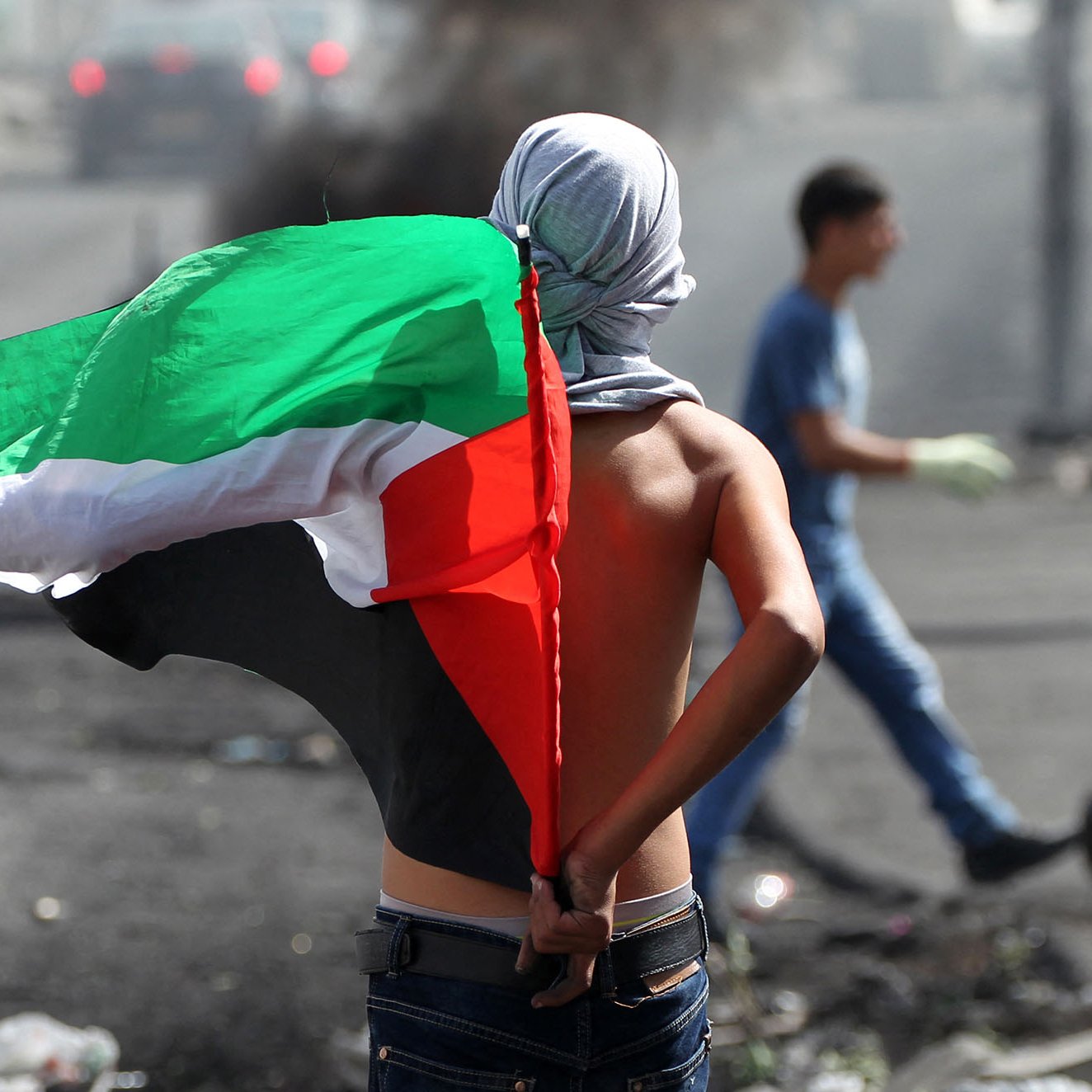 تظهر في الصورة شاب يحمل علم دولة فلسطين، وهو يقف في موقع مظاهرة أو احتجاج. يبدو أنه في وضعية تعبير عن الدعم الوطني، بينما تظهر في الخلفية بعض الدخان أو الفوضى، مما يشير إلى وجود توتر أو صراع في المنطقة. الشاب لا يرتدي قميصًا، مما يعكس أجواء الاحتجاجات أو المظاهرات.