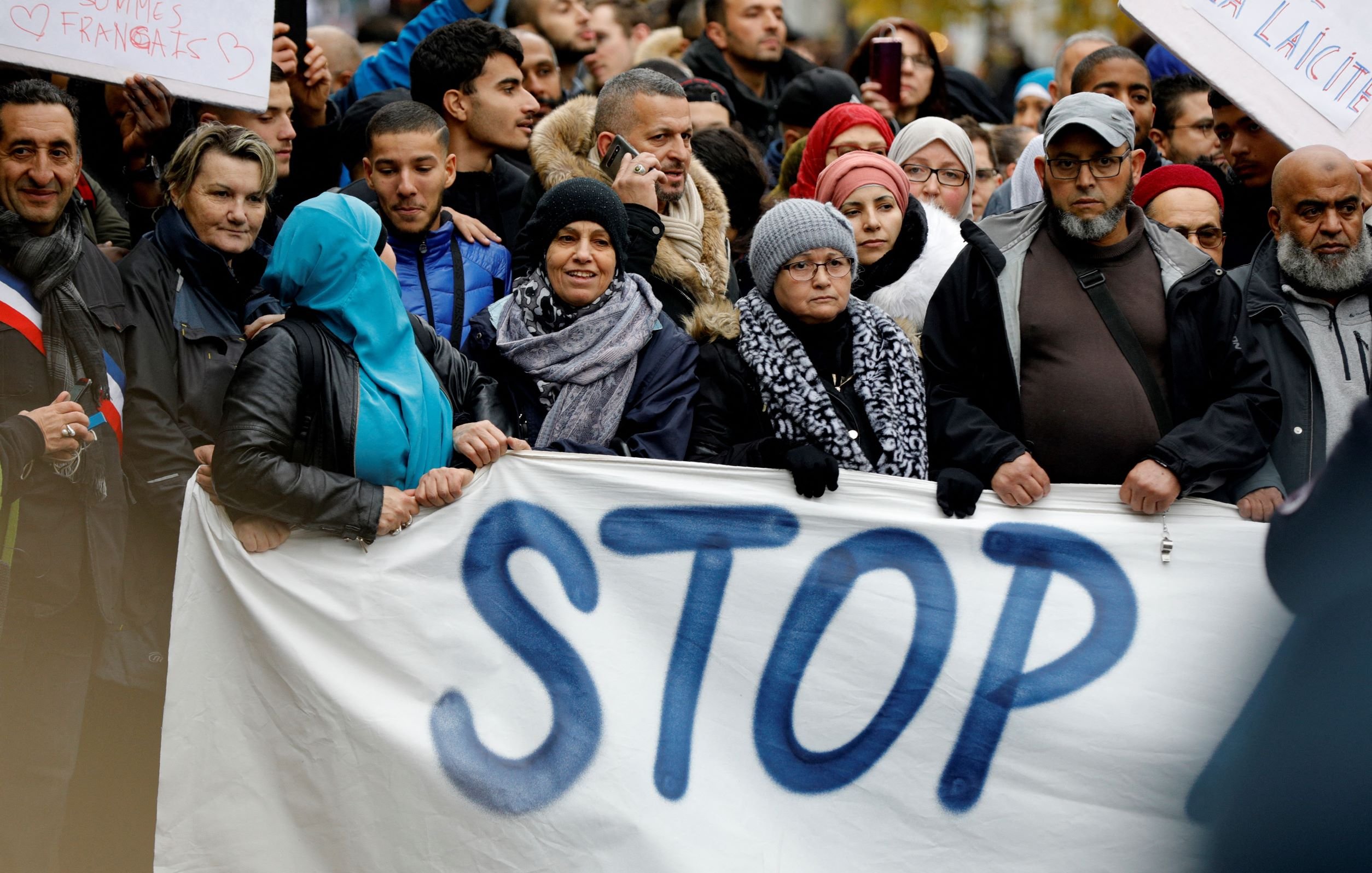 L'image montre une foule de personnes rassemblées pour une manifestation. Elles tiennent des pancartes, dont l'une affiche le mot "STOP" écrit en grandes lettres. Les participants portent des vêtements variés, certains ayant des écharpes ou des chapeaux, ce qui suggère qu'il pourrait faire frais. L'atmosphère semble engagée, avec des visages sérieux ou déterminés, reflétant un fort sentiment de solidarité pour la cause qu'ils soutiennent.