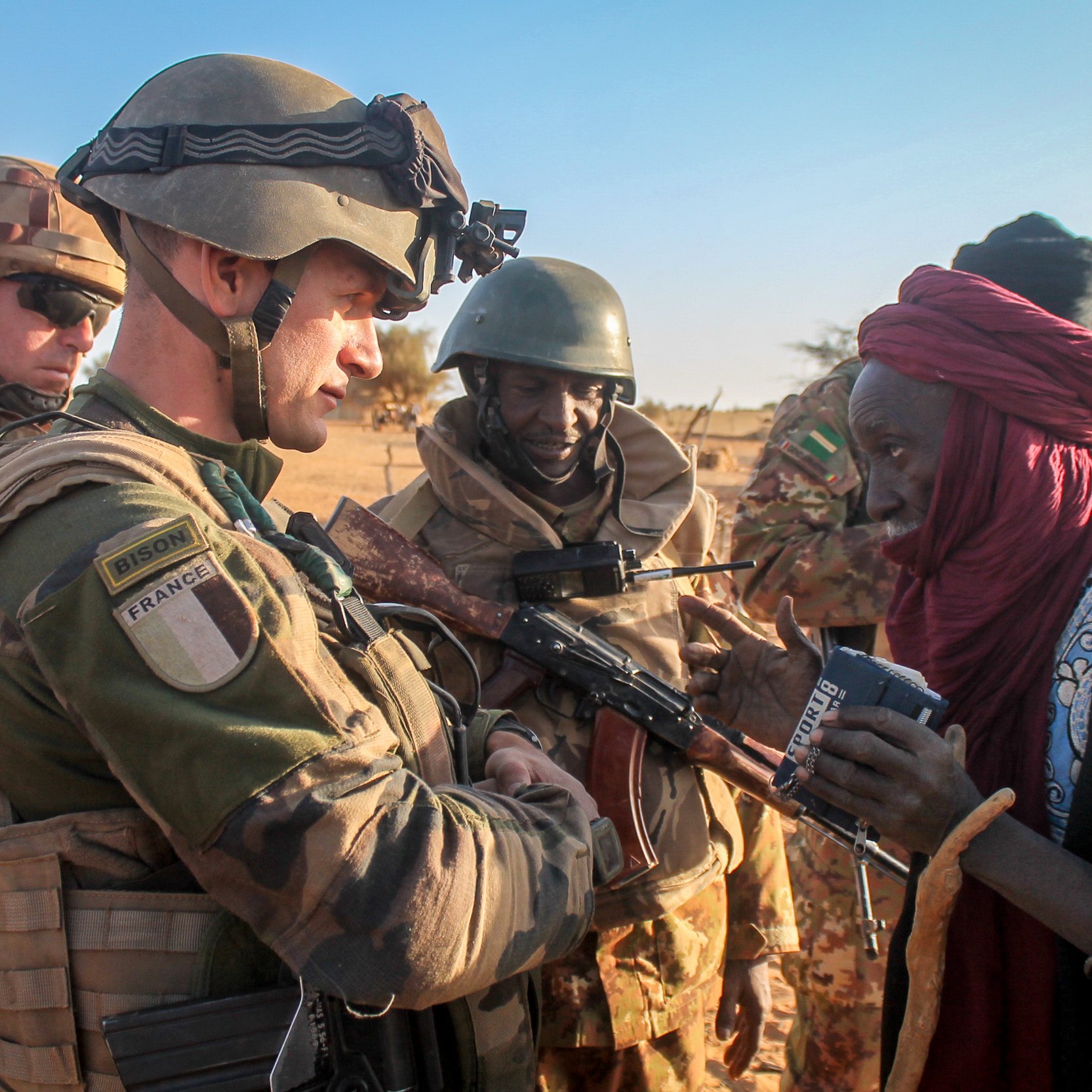 L'image montre un soldat en uniforme militaire, probablement français, interagissant avec un homme âgé, qui semble local. Le soldat est armé et porte un gilet pare-balles ainsi qu'un casque, tandis que l'homme a des vêtements traditionnels. En arrière-plan, d'autres militaires, également en uniforme, observent la scène. Le cadre semble désertique, typique d'une région saharienne, avec un ciel dégagé et un sol sablonneux. L'interaction semble empreinte de respect et d'échange culturel.