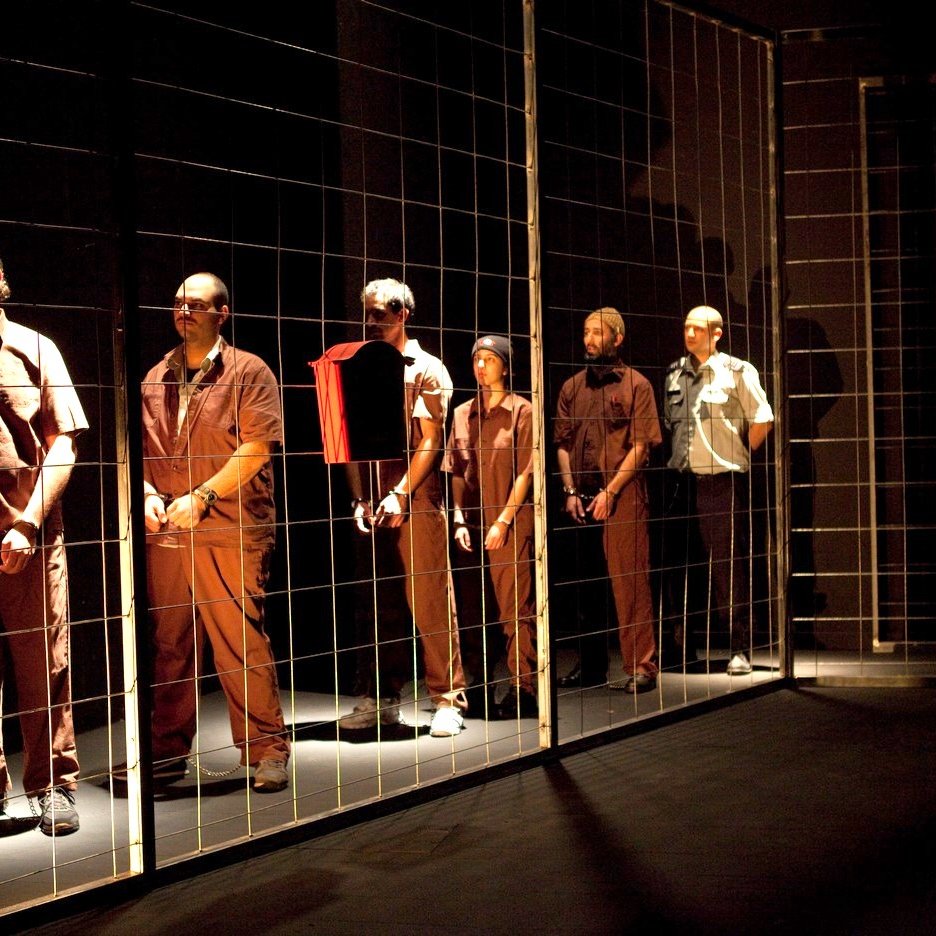 L'image montre un groupe d'hommes disposés en ligne, chacun portant des vêtements de style uniforme. Ils se tiennent derrière un grillage, ce qui crée une impression de séparation ou de détention. L'éclairage est sombre, mettant en valeur les figures des hommes, qui semblent concentrés ou pensifs. L'ambiance générale évoque des thèmes de confinement et d'isolement.