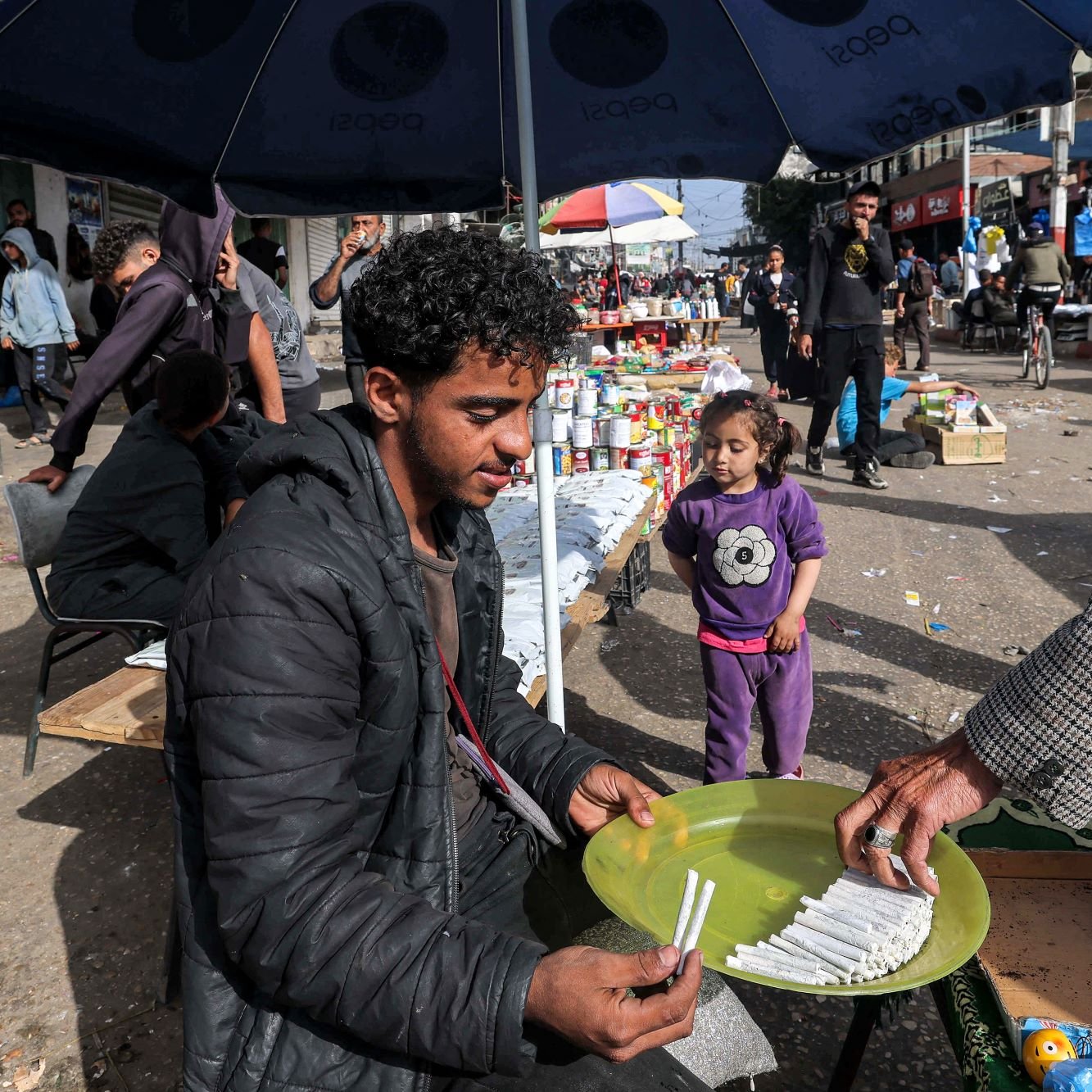 در تصویر، یک بازار محلی به چشم می‌خورد. در وسط تصویر، مرد جوانی نشسته و در حال فروش کالاهایی است. او یک بشقاب سبز رنگ را در دست دارد که در آن چند قلم وجود دارد. به نظر می‌رسد که او به مشتری‌اش توجه دارد که در حال انتخاب کالاهاست. در کنار آن، یک دختر بچه نیز در حال بازی کرده و به صحنه نگاه می‌کند. در پس‌زمینه، افراد دیگری در حال خرید و گشت و گذار هستند و جوی شلوغ و پرجنب‌وجوش به چشم می‌خورد. سایه چترهایی که بالای سر برخی افراد است، فضایی دلپذیر ایجاد کرده است.