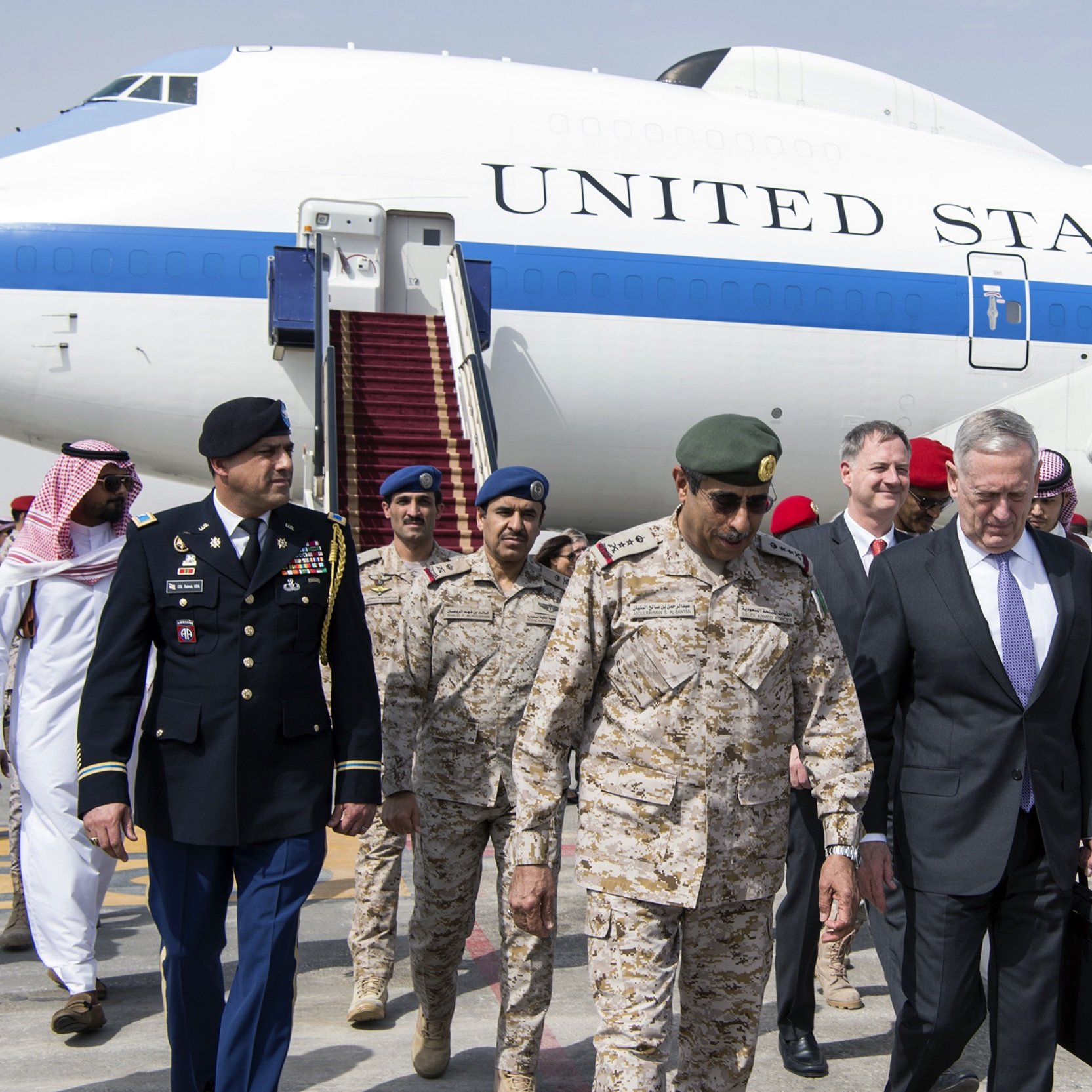 The image depicts a group of individuals exiting from a large aircraft marked with the words "UNITED STATES OF AMERICA." In the foreground, there are several military personnel and officials. One individual appears to be dressed in a military uniform, while others are in formal attire. They seem to be engaged in conversation as they walk down the steps from the plane. The backdrop includes the aircraft and an airport setting, suggesting a diplomatic visit or military engagement.