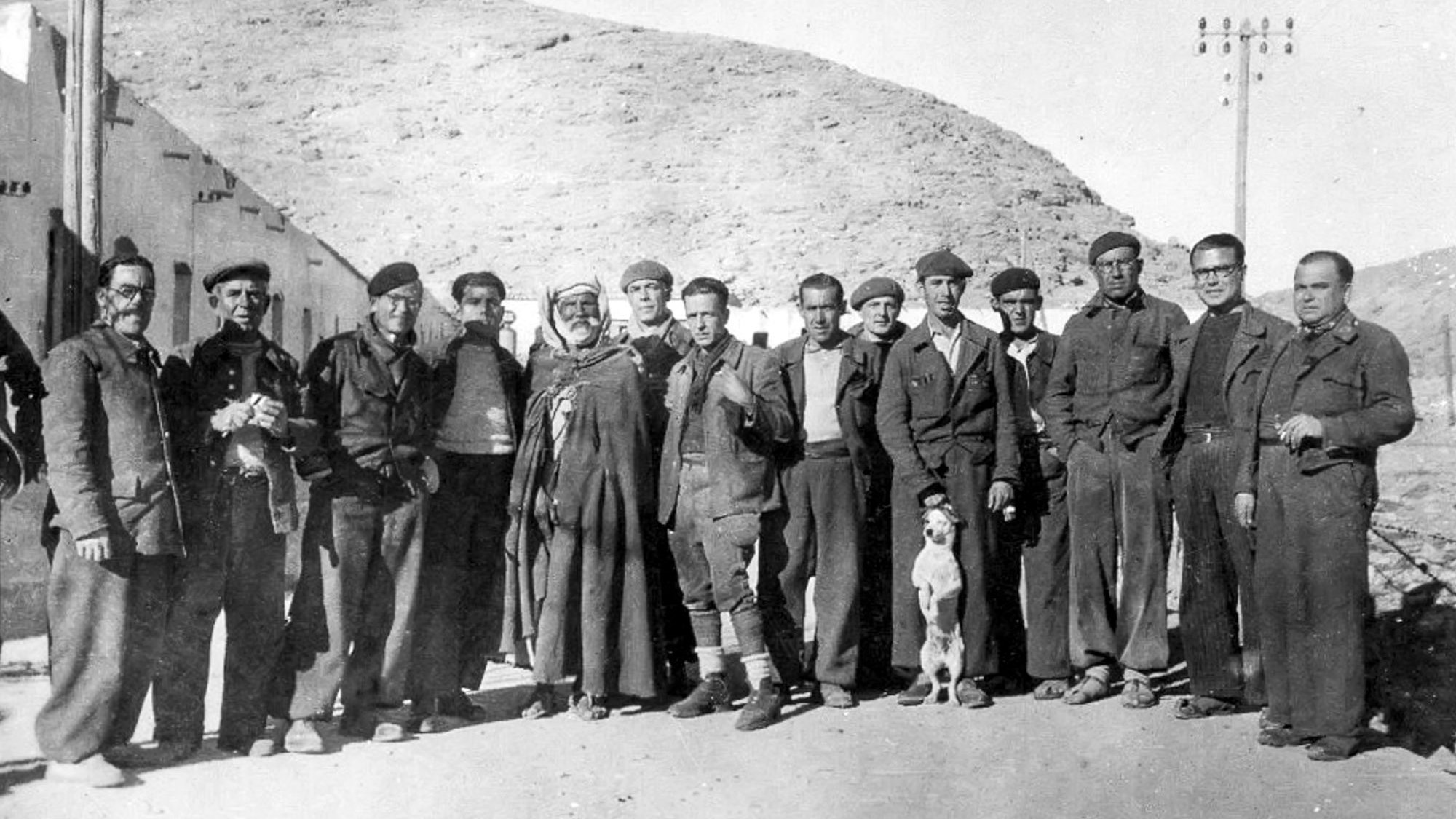 La imagen muestra un grupo de hombres de pie en un entorno rural, posiblemente en una zona montañosa. Están vestidos con ropas tradicionales y de trabajo, algunos con boinas. Hay también un perro que se encuentra entre ellos. La atmósfera es de camaradería, y el paisaje detrás sugiere una época pasada, posiblemente de principios del siglo XX. Los rostros reflejan diferentes expresiones, mientras que la composición transmite una sensación de unión entre los miembros del grupo.