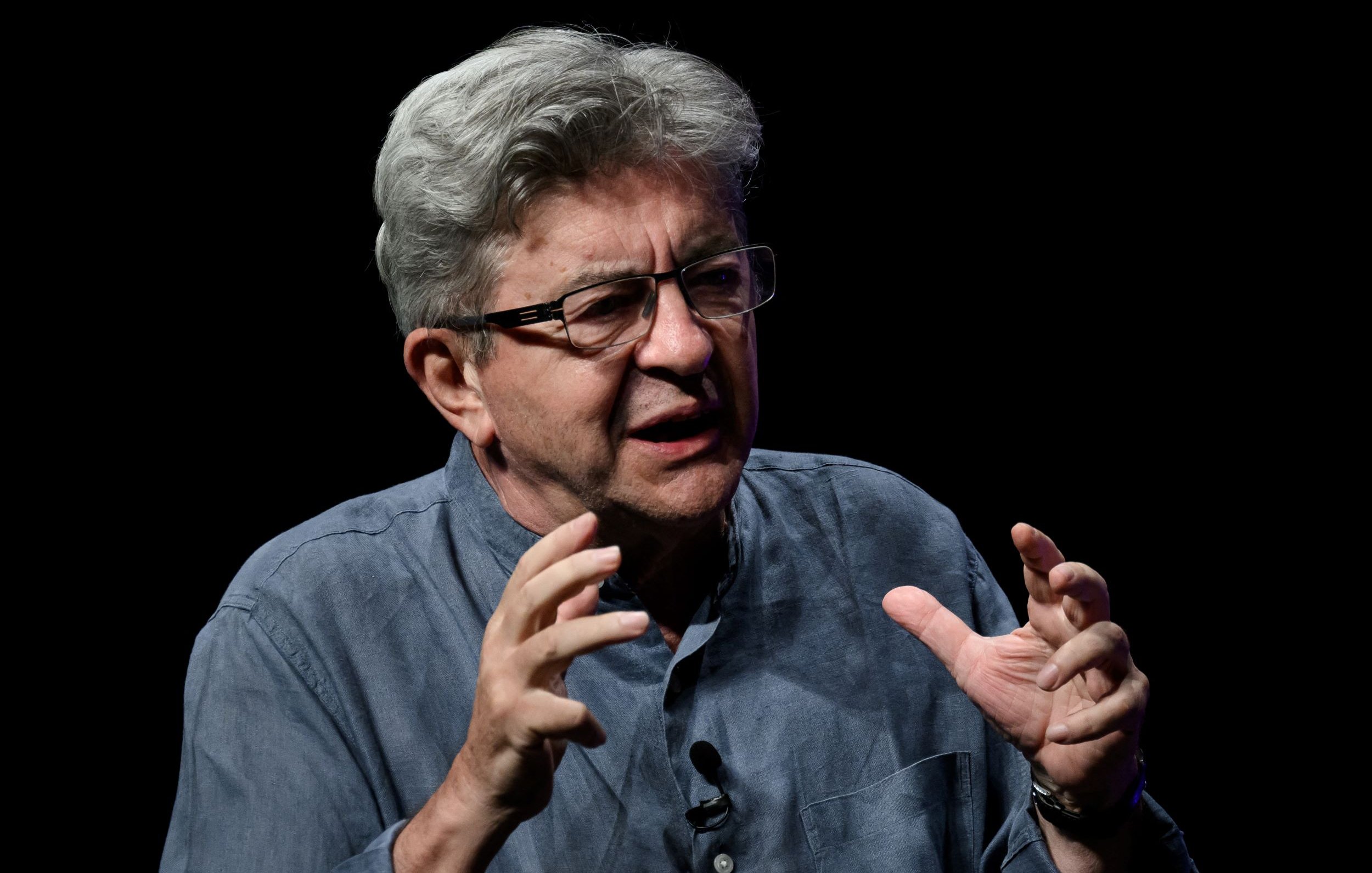 L'immagine mostra un uomo con i capelli grigi, che indossa occhiali e una camicia blu. È impegnato in un discorso appassionato, con le mani sollevate in un gesto espressivo. Sullo sfondo, c'è un'illuminazione scura che mette in risalto il suo viso e la sua gestualità. La sua espressione sembra indicare un forte coinvolgimento nel tema di cui sta parlando.