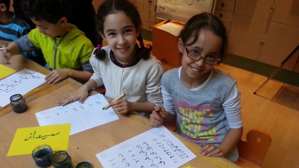 L'image montre deux jeunes filles assises à une table, souriant et travaillant sur des feuilles de papier. Elles utilisent des pinceaux et de l'encre, et semblent engagées dans une activité créative, probablement de calligraphie. Des étiquettes jaunes sont visibles, sur l'une est écrit "سيماء" et sur l'autre "دنيا" en arabe. L'environnement est scolaire, avec d'autres enfants visibles en arrière-plan.