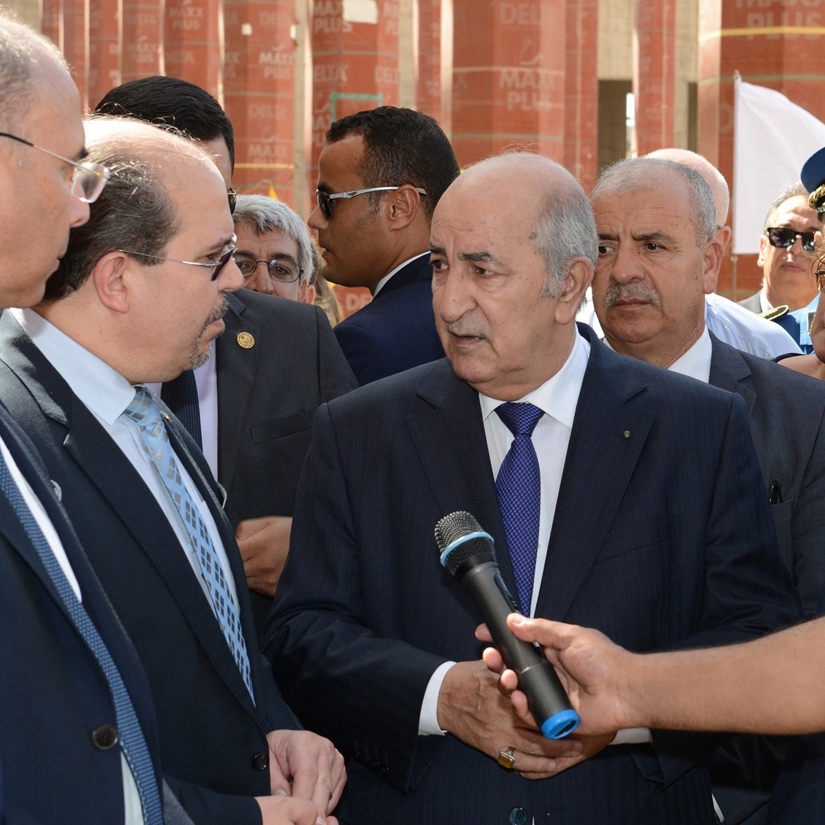 L'image montre un groupe d'hommes en costume, probablement en train de discuter. Au centre, l'un des hommes semble occuper une position de leader, tandis que les autres l'écoutent attentivement. Ils semblent être dans un cadre formel, peut-être lors d'une visite officielle ou d'un événement important. L'environnement en arrière-plan suggère un lieu institutionnel, avec des structures architecturales visibles.